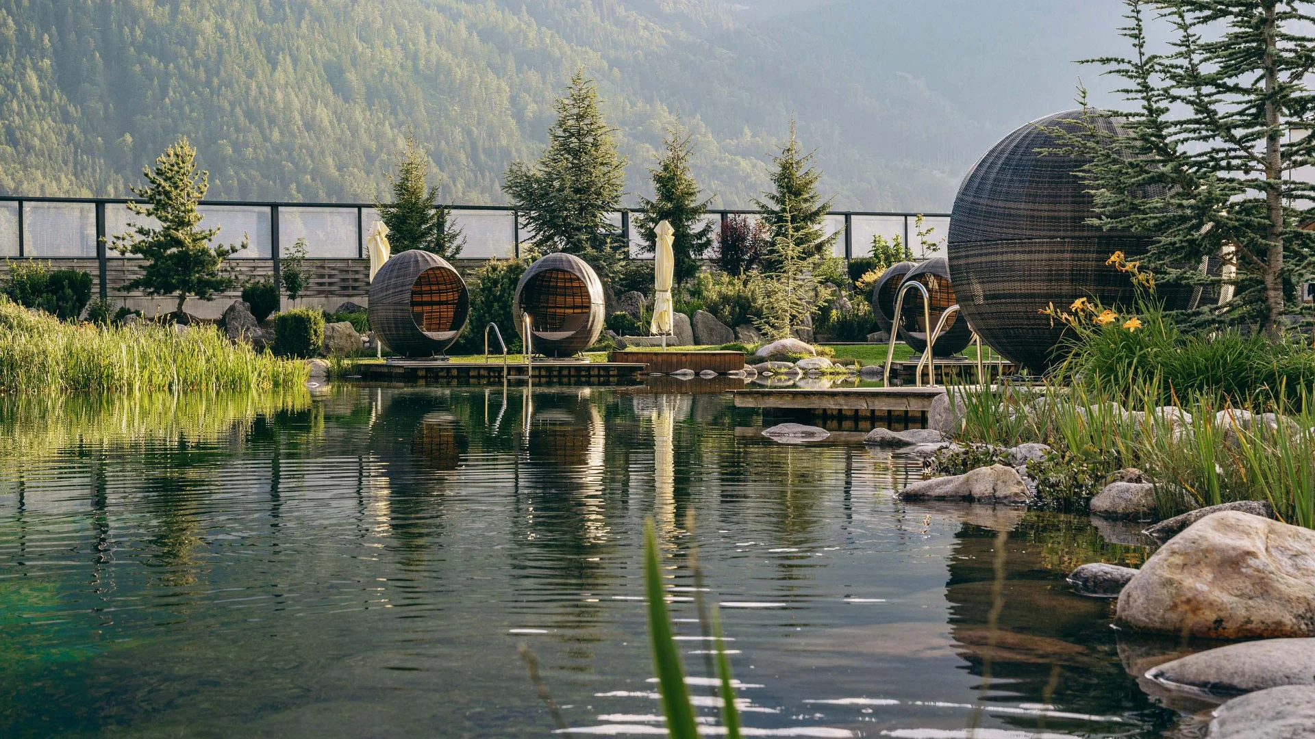 Welcome to our family hotel at Lake Achensee. Natural pool with round seating pods in a garden area with mountains in the background
