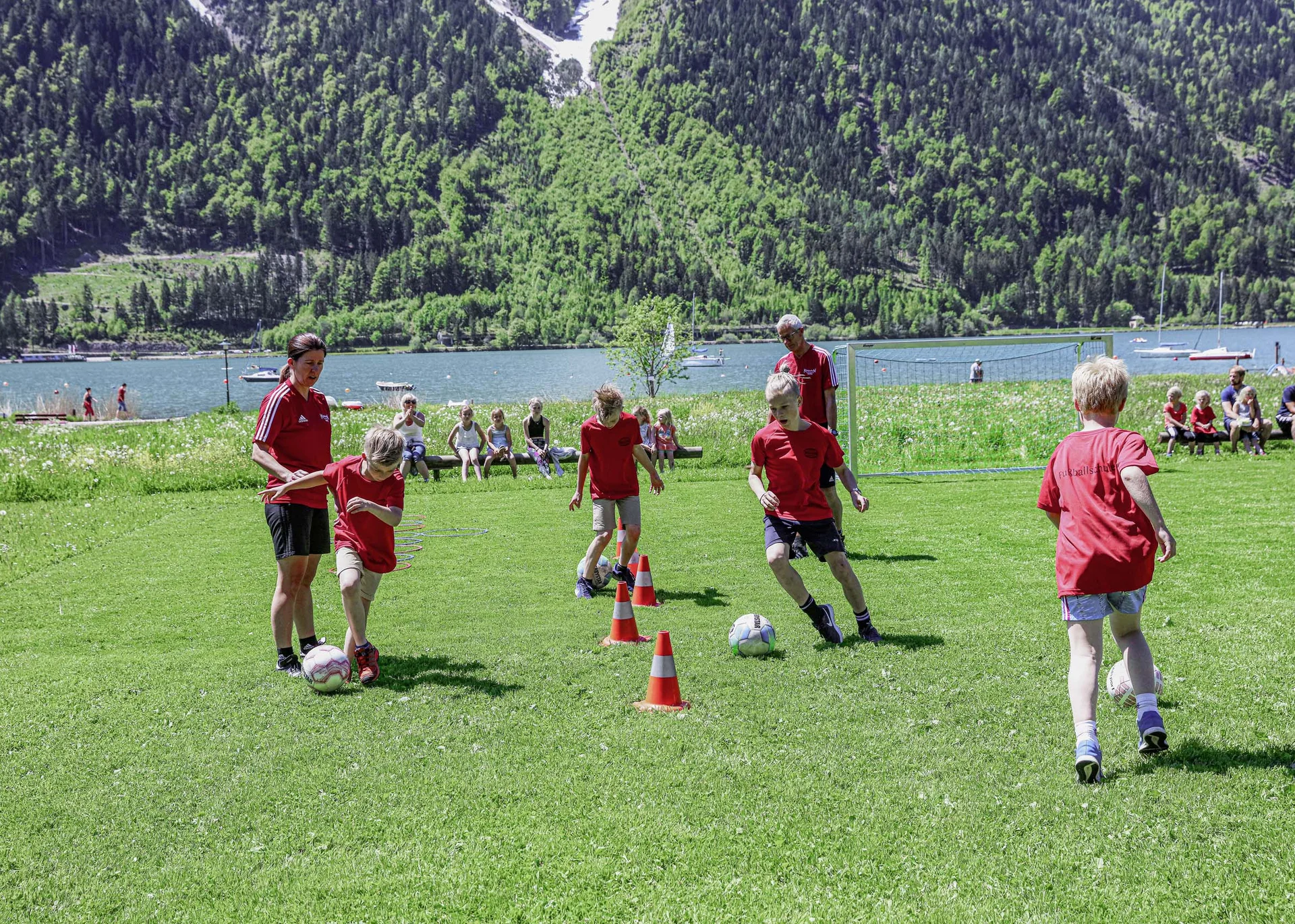 Aktiver Urlaub im Hotel mit Fußballcamp Kinder trainieren Fußball mit Trainern auf Wiese mit Bergen und See im Hintergrund