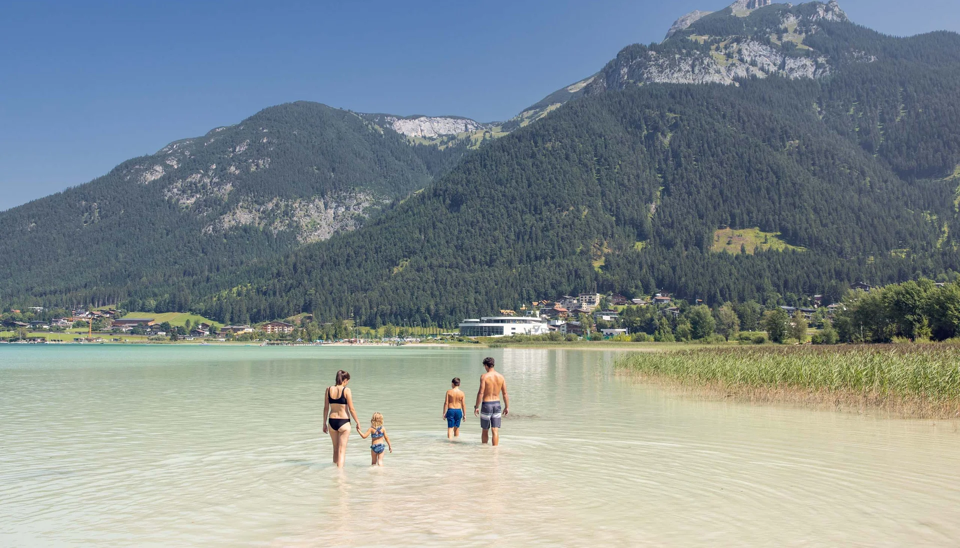 Familienzeit im Kinderhotel Buchau am Achensee Familie watet in klarem Wasser vor Bergkulisse und Dorf im Hintergrund