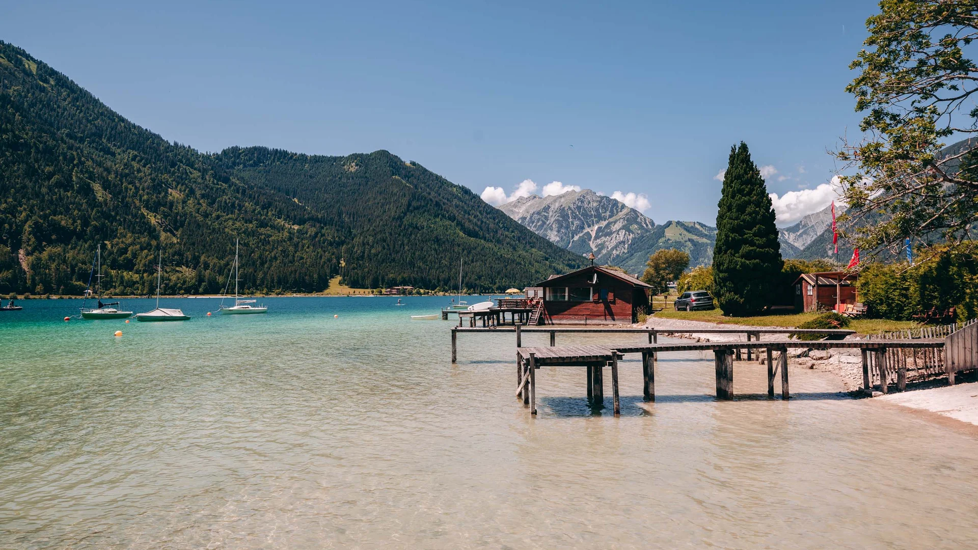 Wassersport auf dem See in Österreich Klares Wasser, Boote und Holzanleger an einem Bergsee mit Hütten und Bergen im Hintergrund