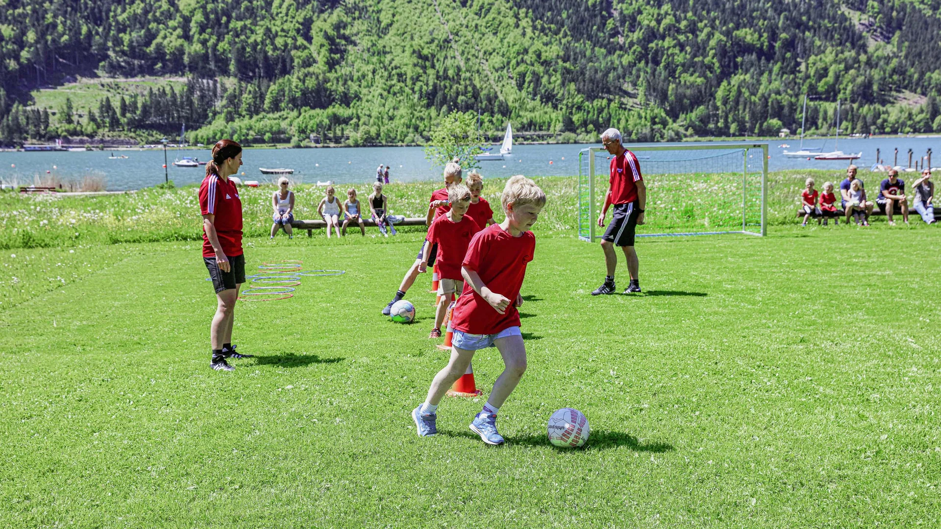 Aktiver Urlaub im Hotel mit Fußballcamp Kinder trainieren Fußball mit Trainern auf einer Wiese am See vor Zuschauern