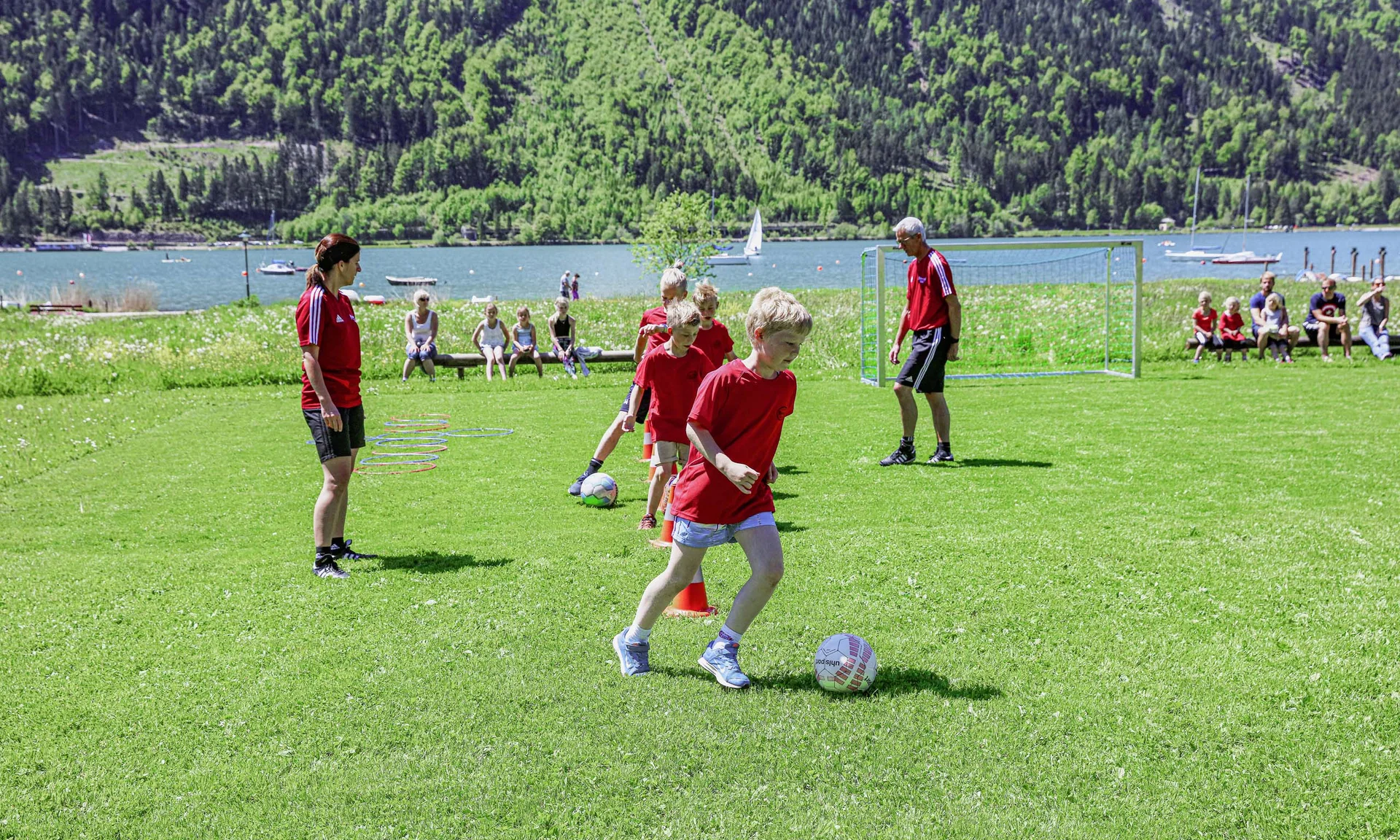 Outdoorspaß im Familienhotel am See in Österreich Kinder trainieren Fußball mit Trainern auf einer Wiese am See vor Zuschauern