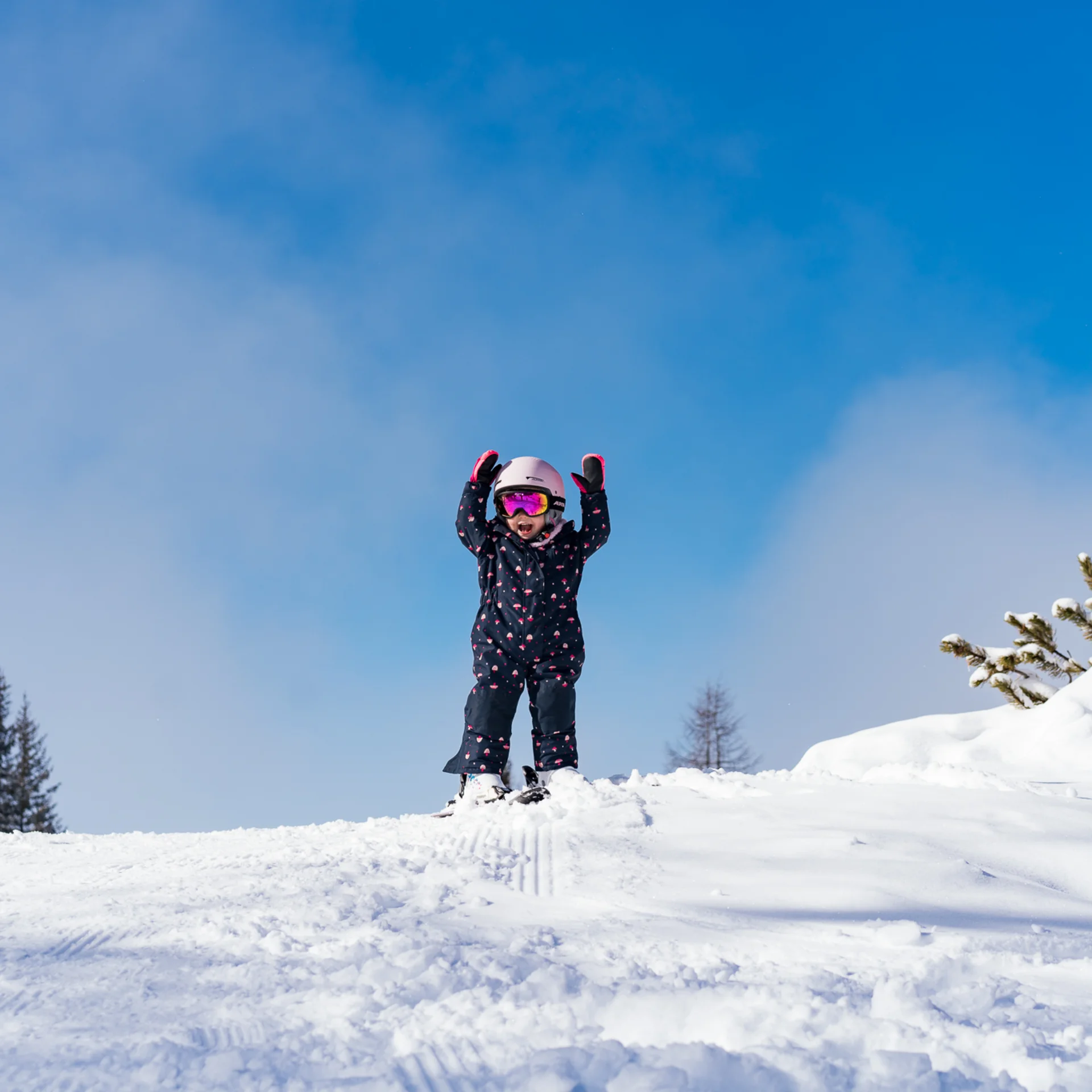 Der Wintertraum im Familien-Skihotel in Österreich Kind in Skikleidung hebt die Arme auf schneebedecktem Hügel bei blauem Himmel
