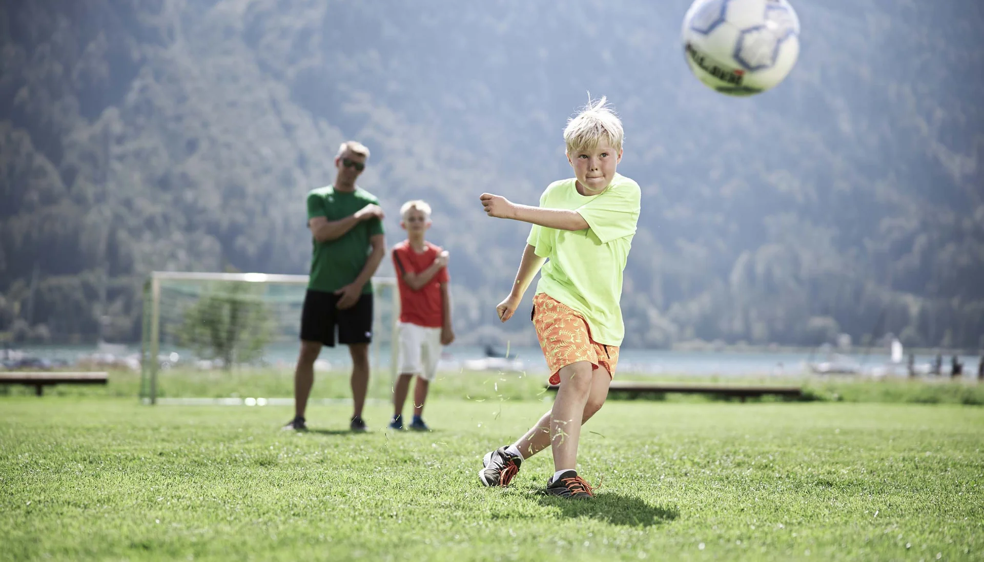 Aktiver Urlaub im Hotel mit Fußballcamp Junge schießt Fußball auf grünem Feld mit zwei Personen im Hintergrund