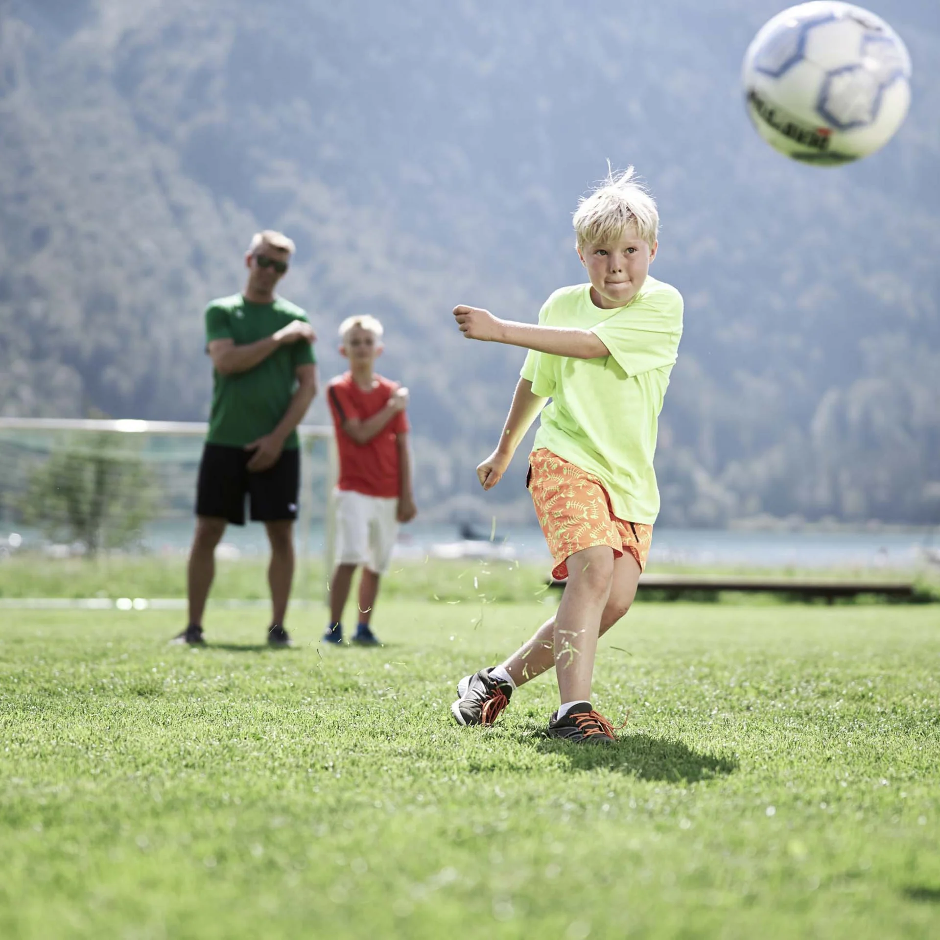 Welcome to our family hotel at Lake Achensee. Boy kicking soccer ball on grass field with two people watching behind