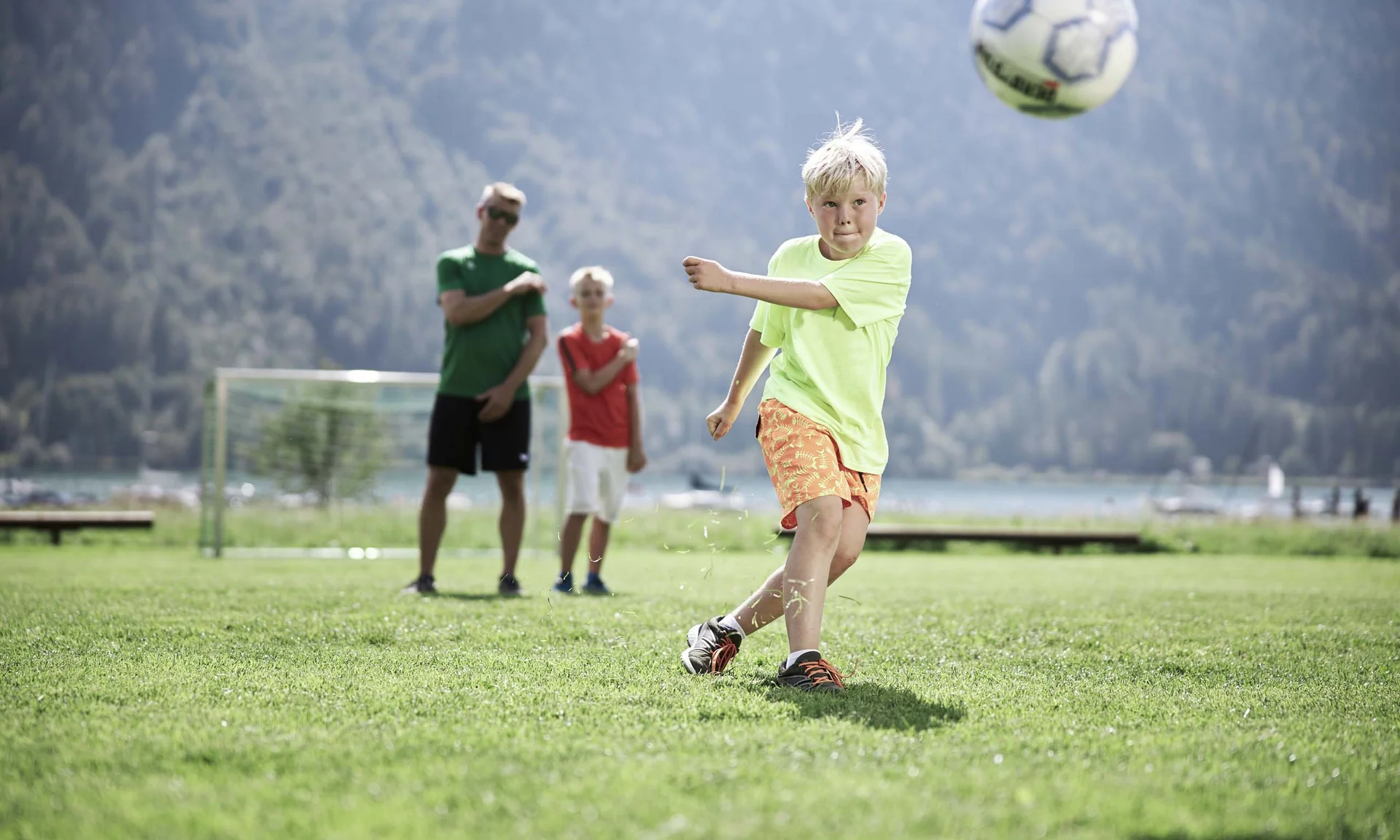 Freizeitkurse für Ihre Kids und Teenies Junge schießt Fußball auf grünem Feld mit zwei Personen im Hintergrund