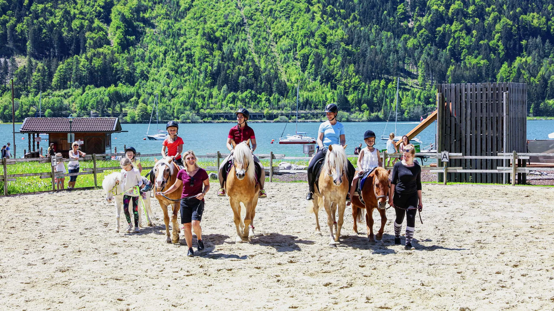 Pferdeglück im Reithotel in Österreich Kinder reiten Pferde in einer sonnigen Arena vor einem See und bewaldeten Hügeln