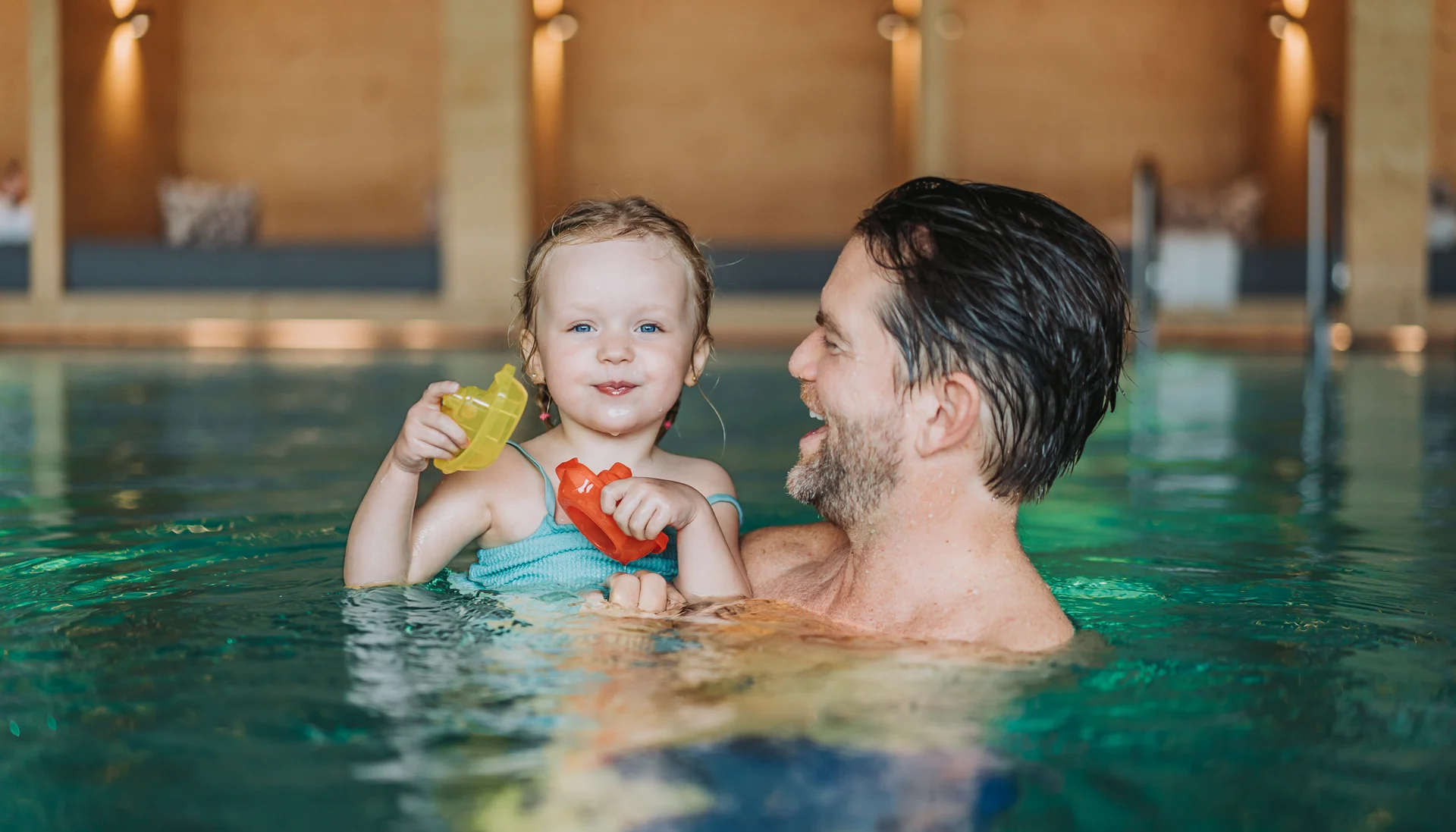 Wasserspaß im Familienhotel in Tirol mit Pool Vater hält lachendes kleines Mädchen mit Spielzeugbooten im Hallenbad