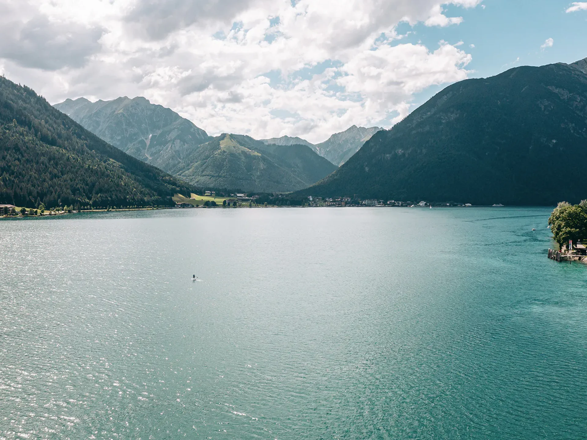 Wassersport auf dem See in Österreich Bergsee mit bewaldeten Bergen, leicht bewölktem Himmel und einer Person auf einem Stand-up-Paddleboard