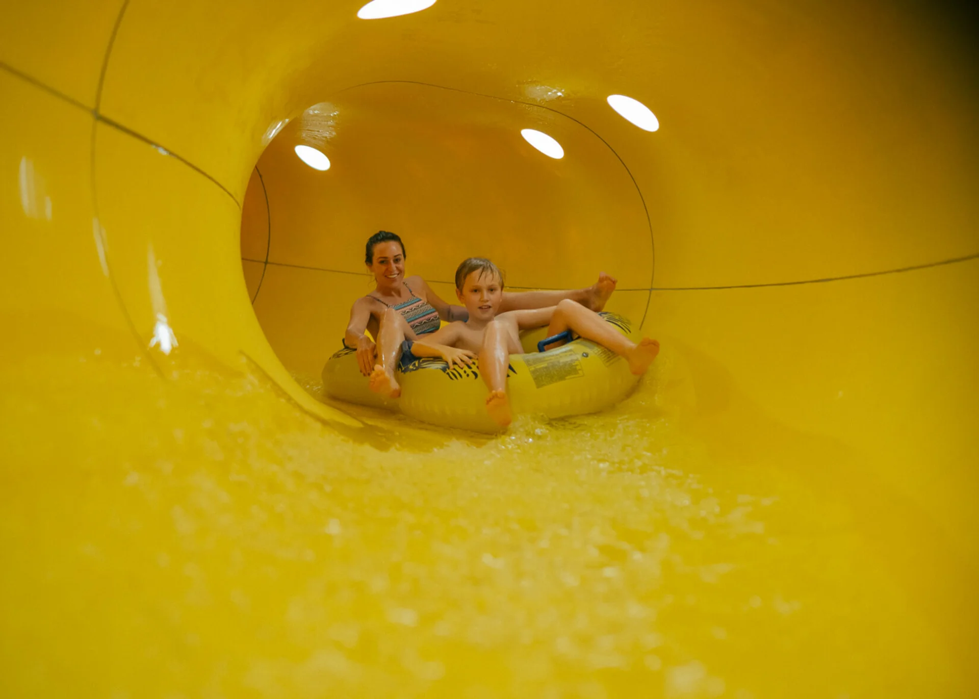 Wasserspaß im Familienhotel in Tirol mit Pool Mutter und Sohn fahren gemeinsam auf einem Schlauchreifen in einer gelben Wasserrutsche