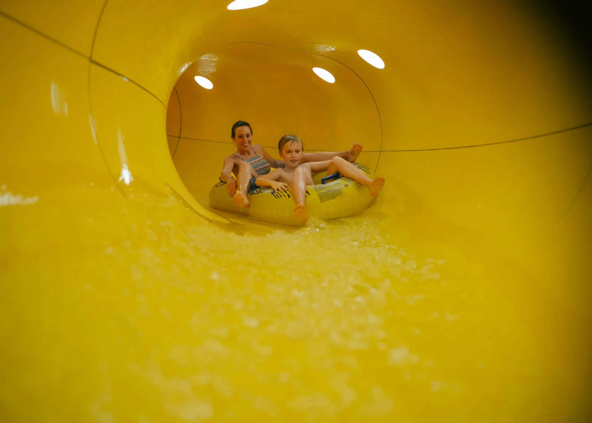 Urlaub im Hotel mit Indoorspielplatz Mutter und Sohn fahren gemeinsam auf einem Schlauchreifen in einer gelben Wasserrutsche