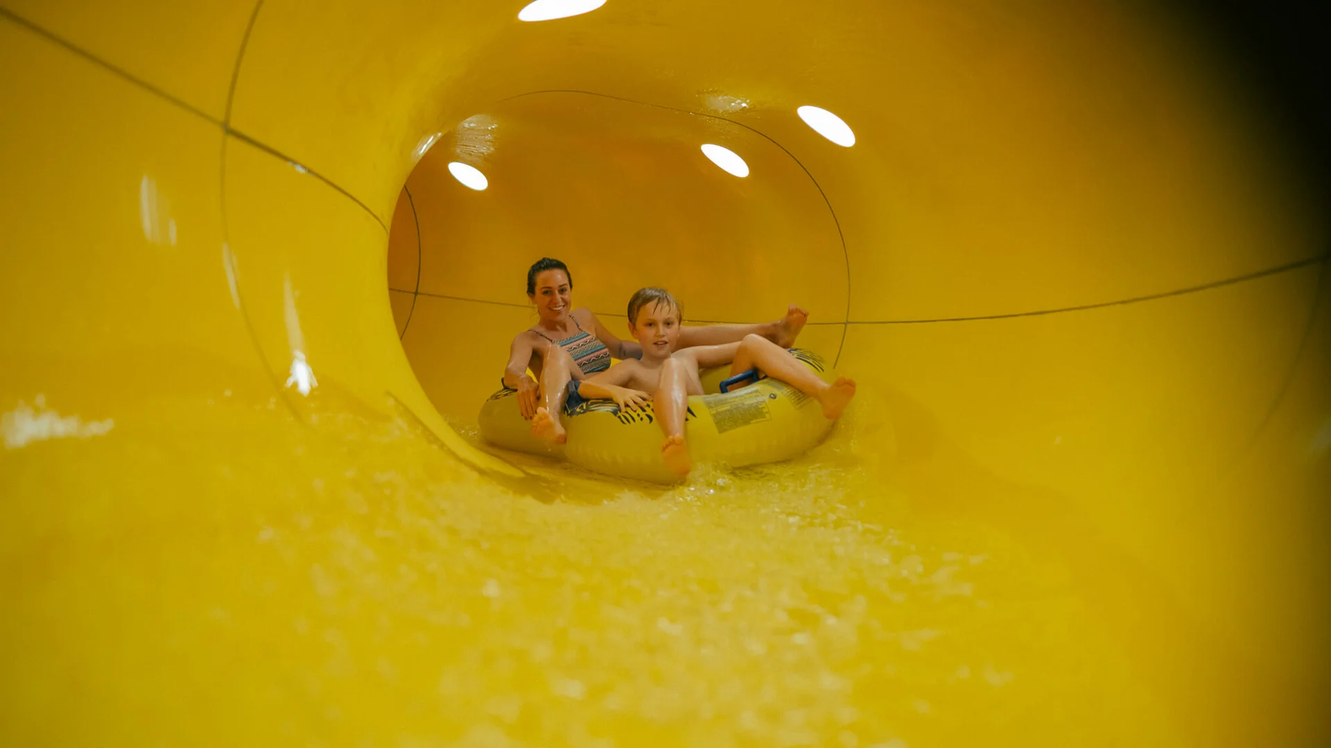 Urlaub im Hotel mit Indoorspielplatz Mutter und Sohn fahren gemeinsam auf einem Schlauchreifen in einer gelben Wasserrutsche