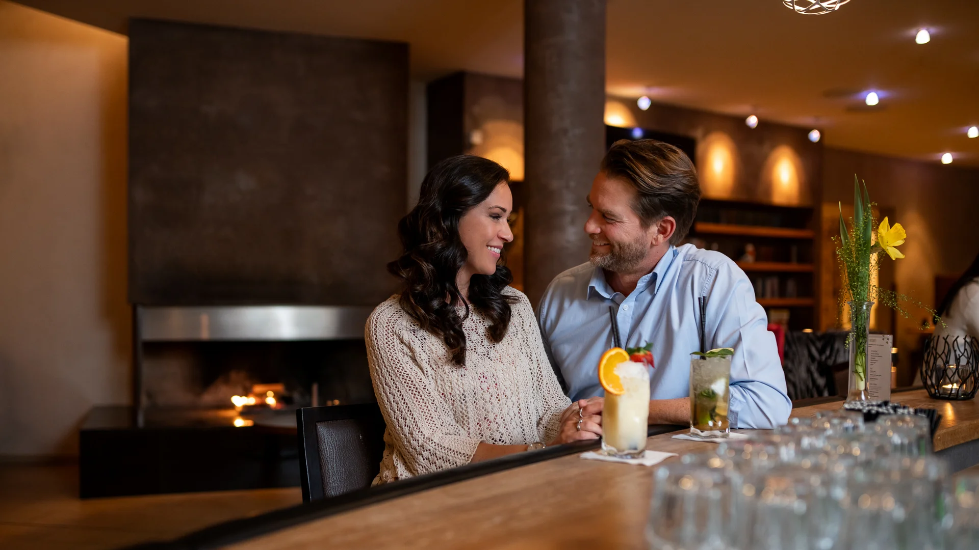 Erholungsurlaub für Eltern Paar sitzt an Bar mit Cocktails in gemütlicher Bar mit Kamin im Hintergrund