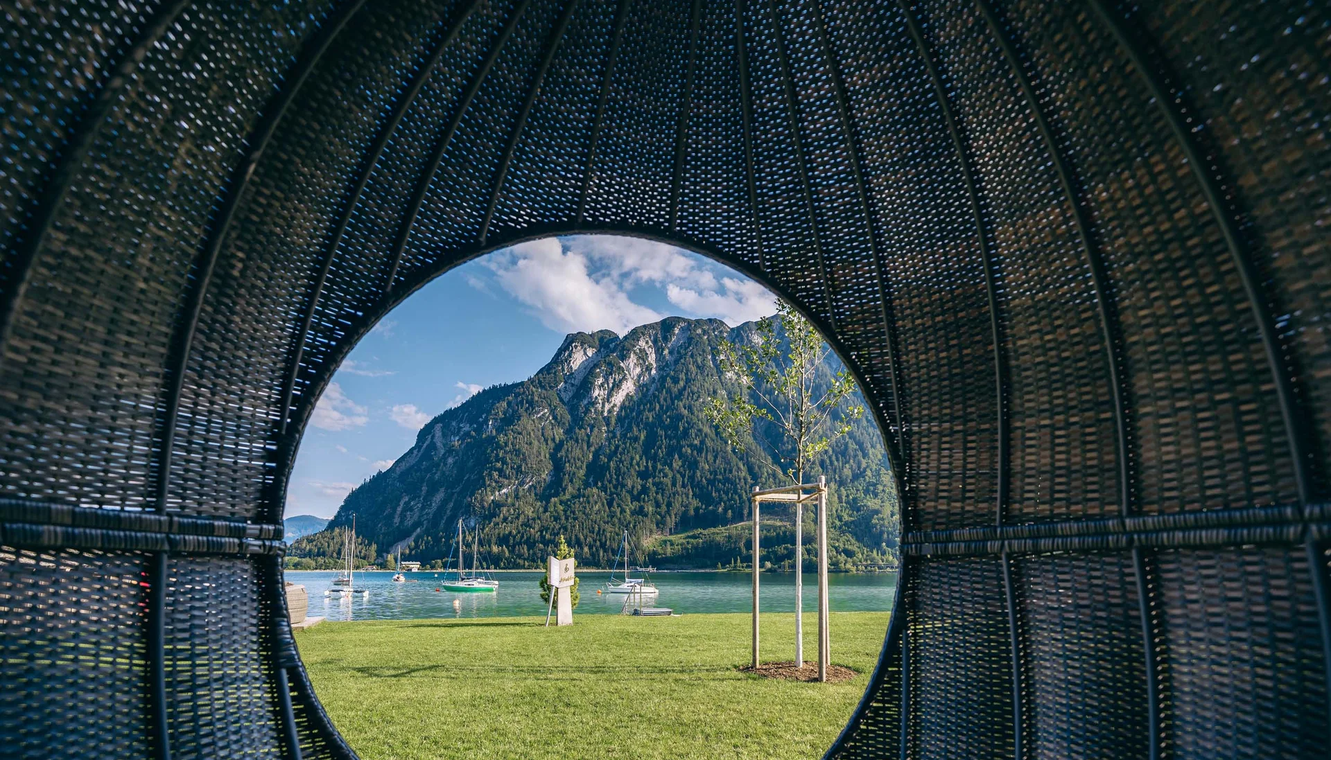 Wassersport auf dem See in Österreich Blick durch ein rundes Korbgewebe auf Berg, See und Wiese mit Segelbooten