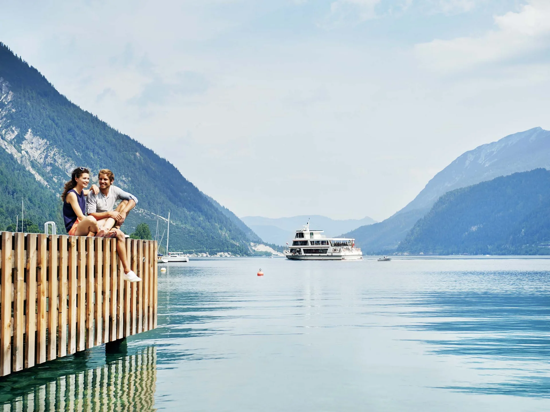 Erholungsurlaub für Eltern Paar sitzt am Steg eines Sees mit Berglandschaft und Ausflugsboot im Hintergrund