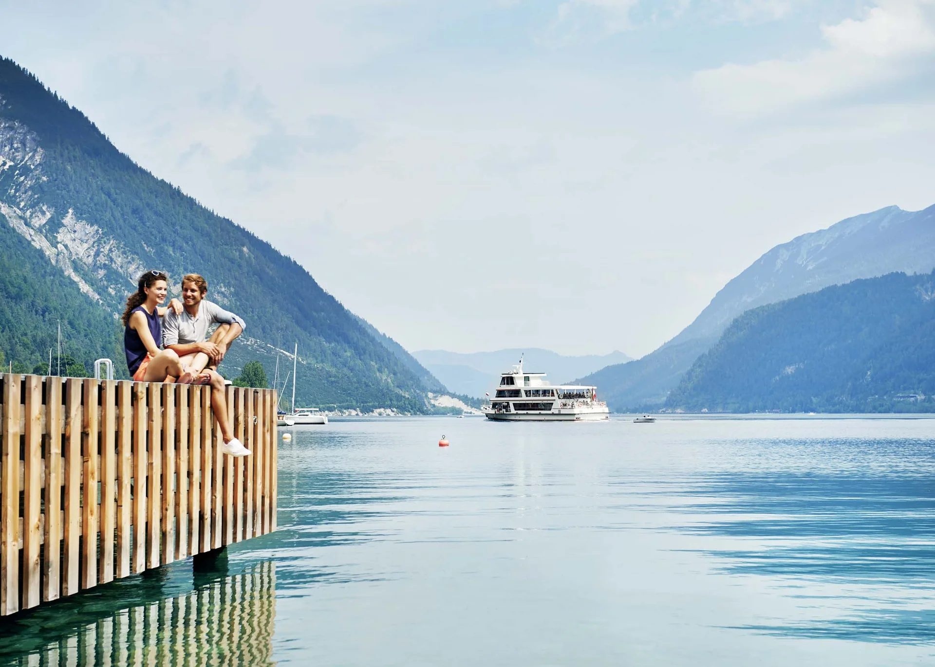Your unforgettable active lakeside holiday in Austria Couple sitting on a dock by a lake with mountains and a tour boat in the background