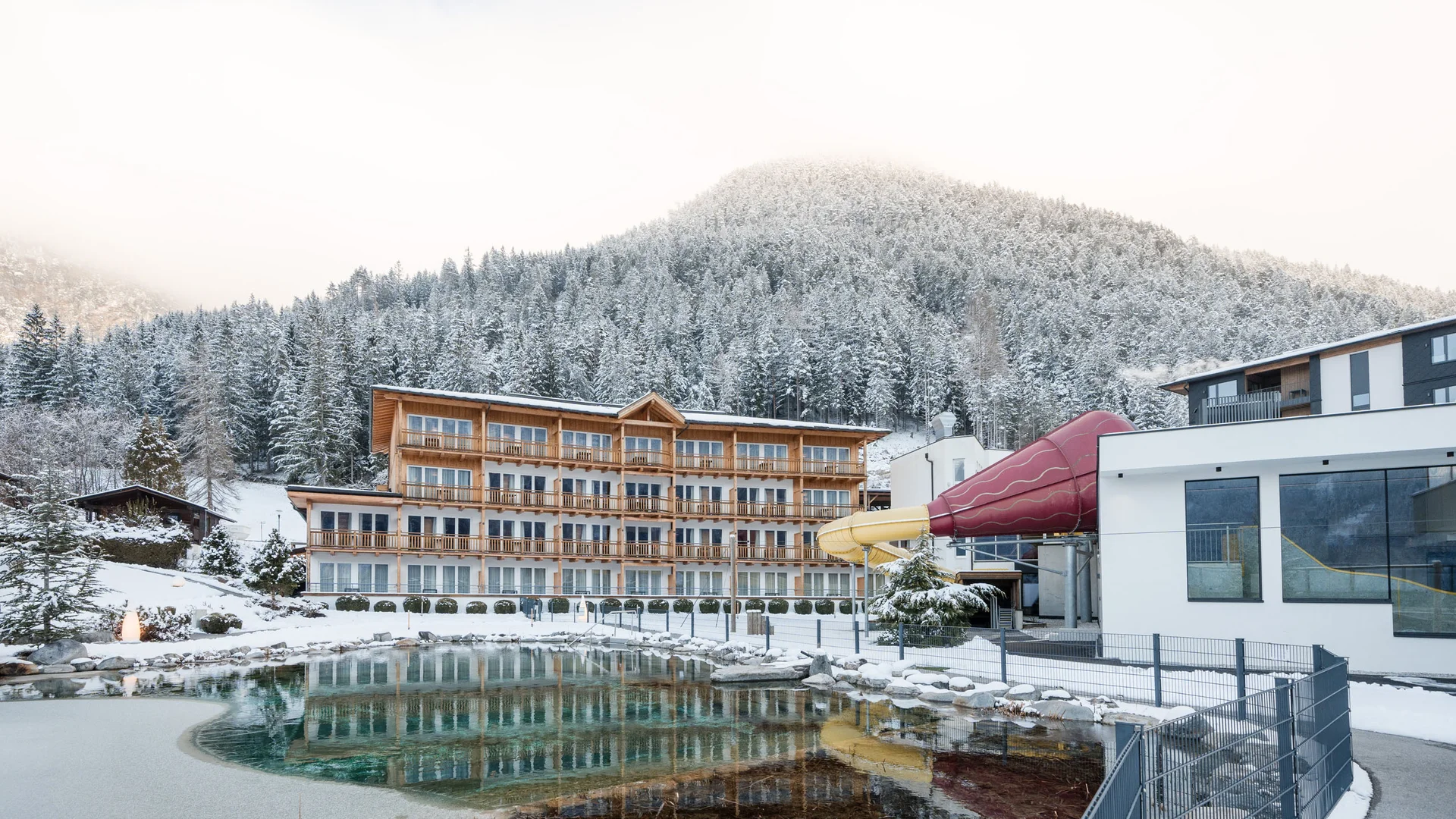 Willkommen im Familienhotel Buchau am Achensee Hotel mit Rutsche und Teich vor verschneitem Berg im Winter