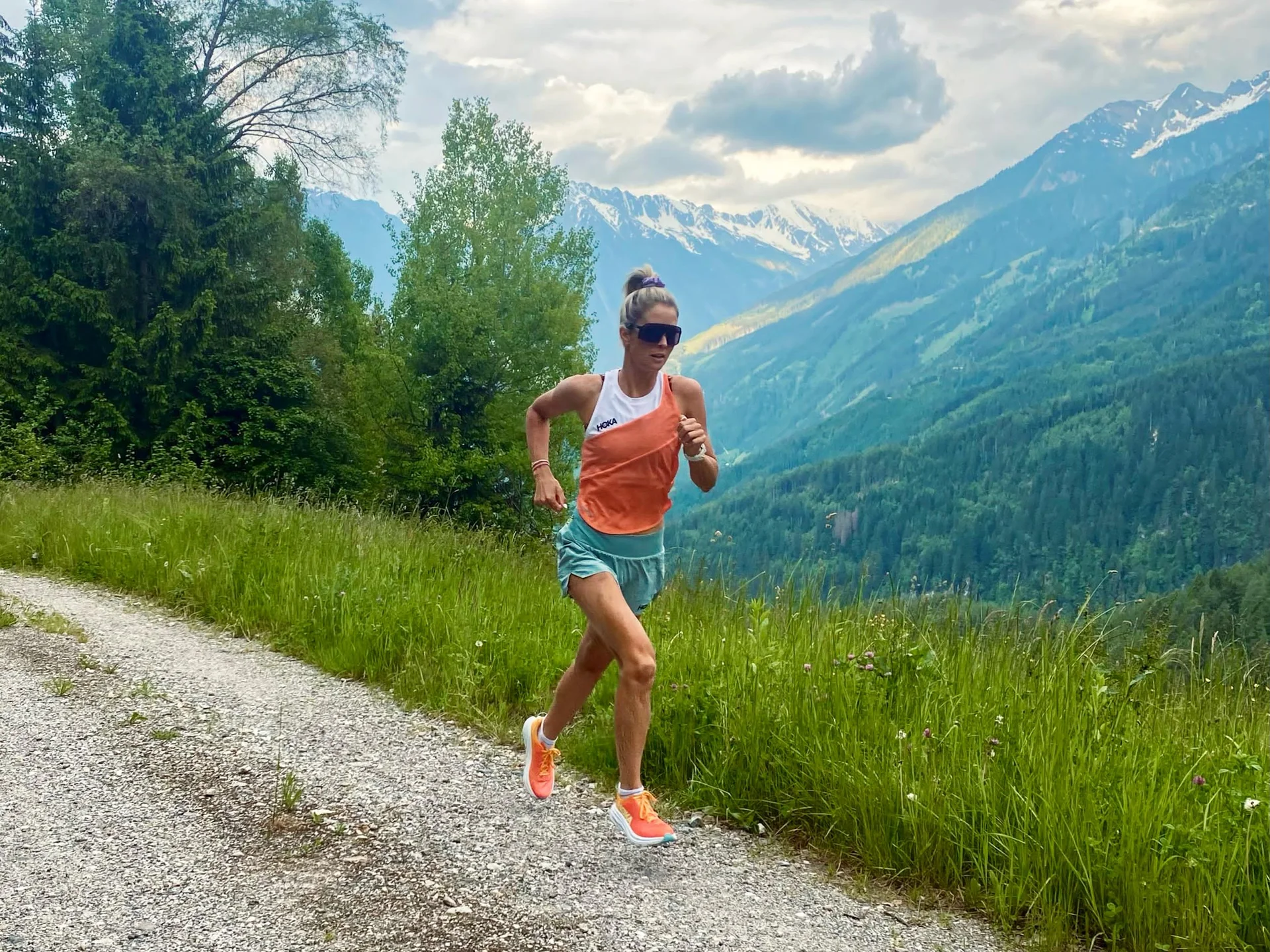 Fitness und Yoga im Hotel in Tirol Frau läuft auf einem Bergweg mit grünen Bäumen und schneebedeckten Bergen im Hintergrund