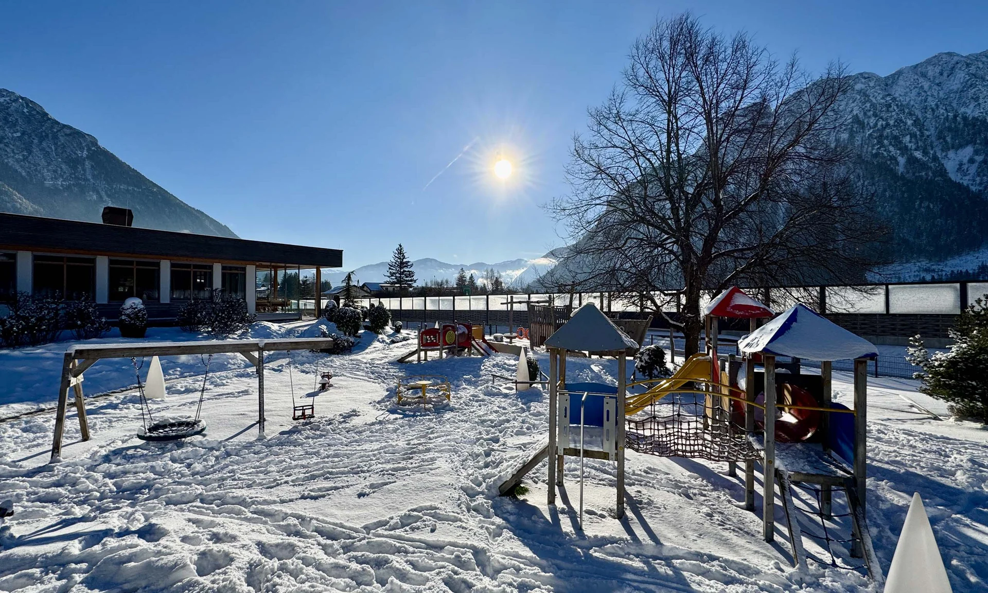 Wir lassen Bilder sprechen. Schneebedeckter Kinderspielplatz mit Rutschen, Schaukel und Bergen im Hintergrund