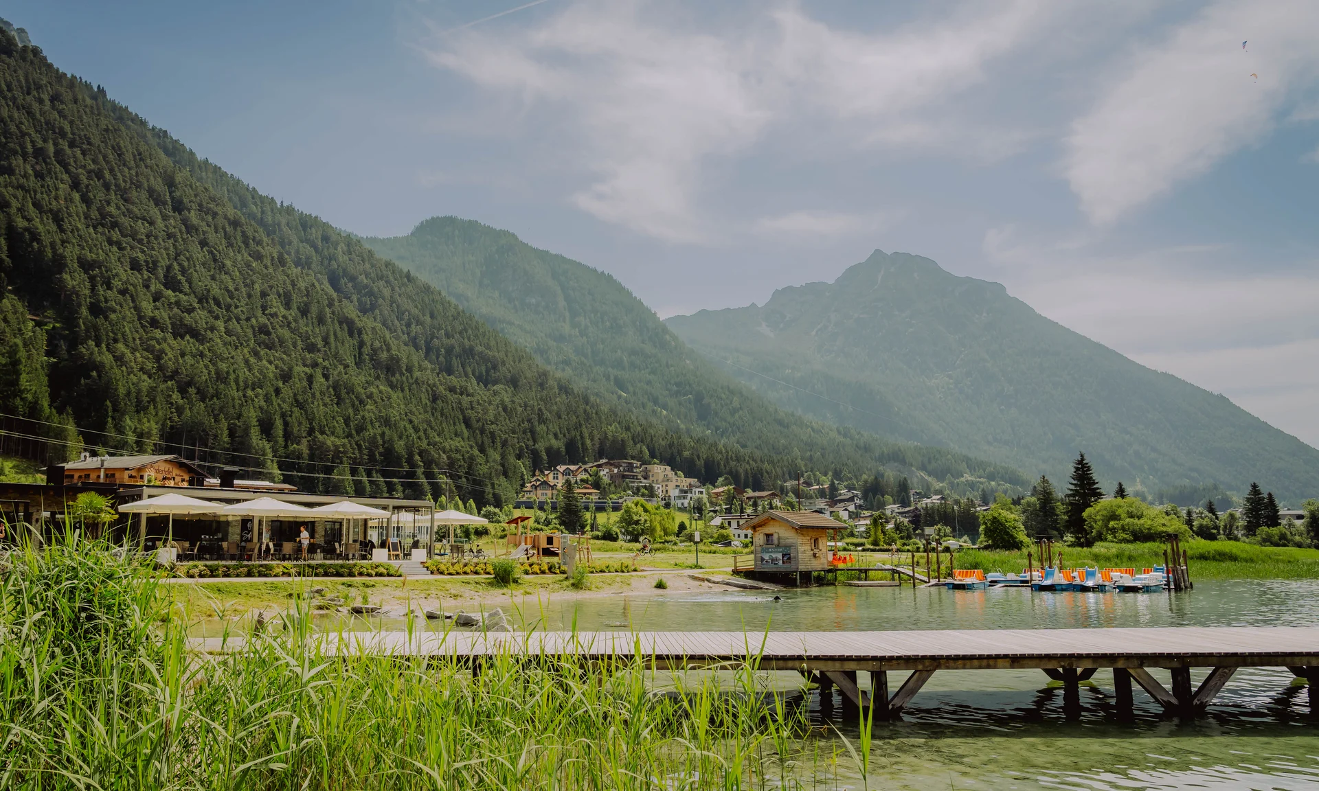 Wir lassen Bilder sprechen. Steg an einem Bergsee mit Booten, Häusern und bewaldeten Bergen im Hintergrund