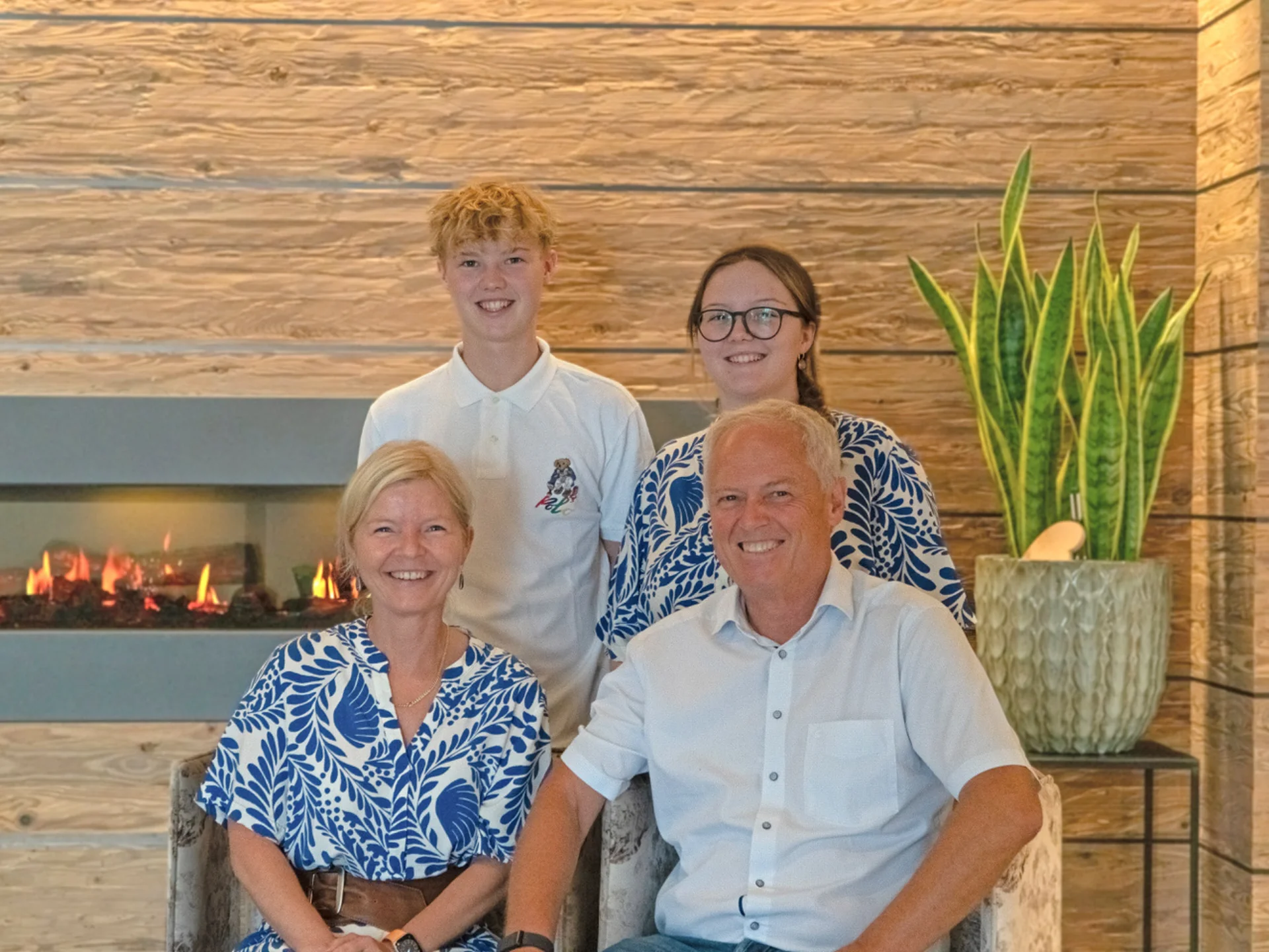 Urlaub im Familienresort in Österreich Fröhliche Familie mit zwei Kindern vor einem Kamin und einer Pflanze im Wohnzimmer