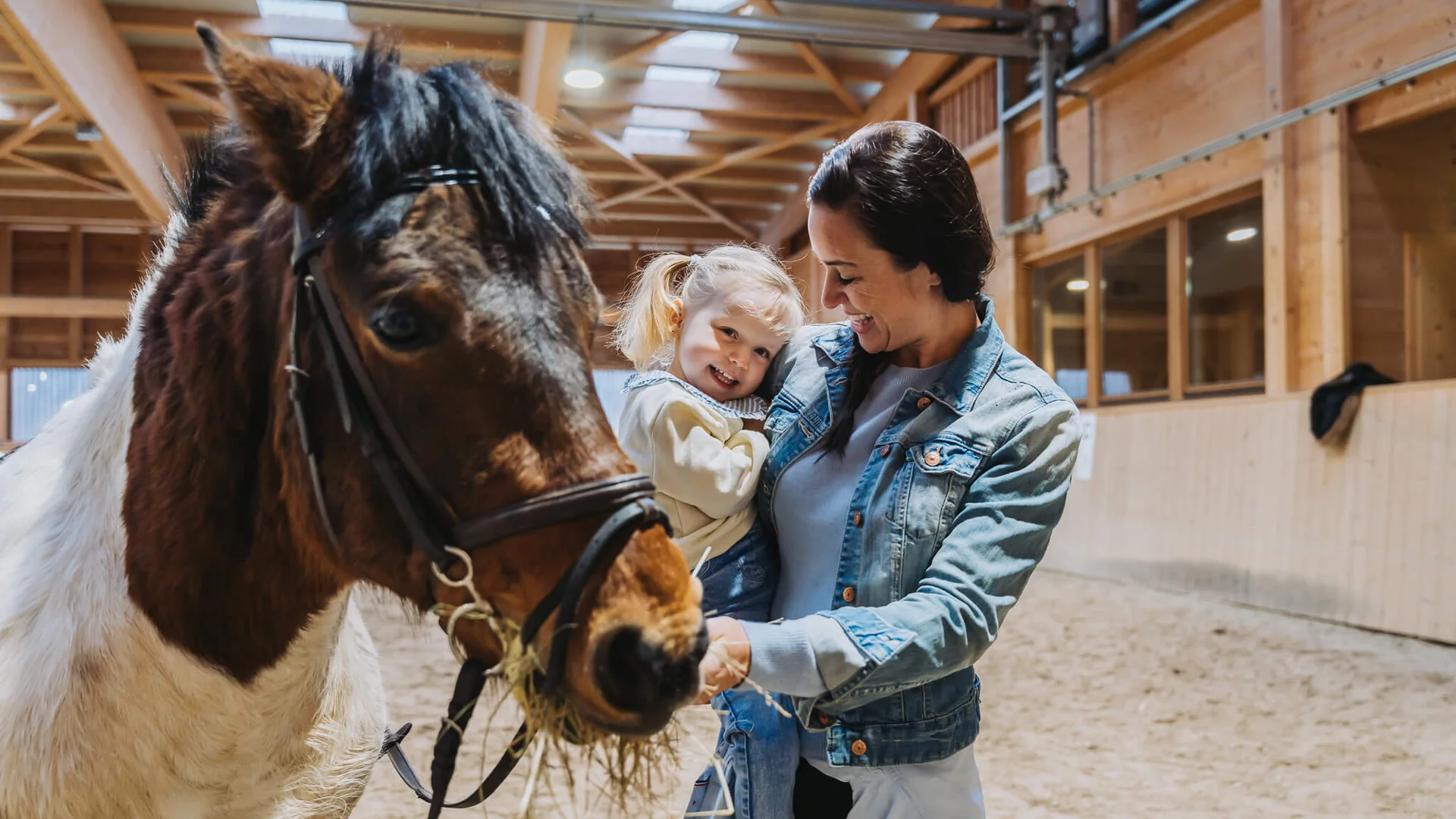 Pferdeglück im Reithotel in Österreich Frau hält lachendes Mädchen im Innenstall neben fressendem Pony