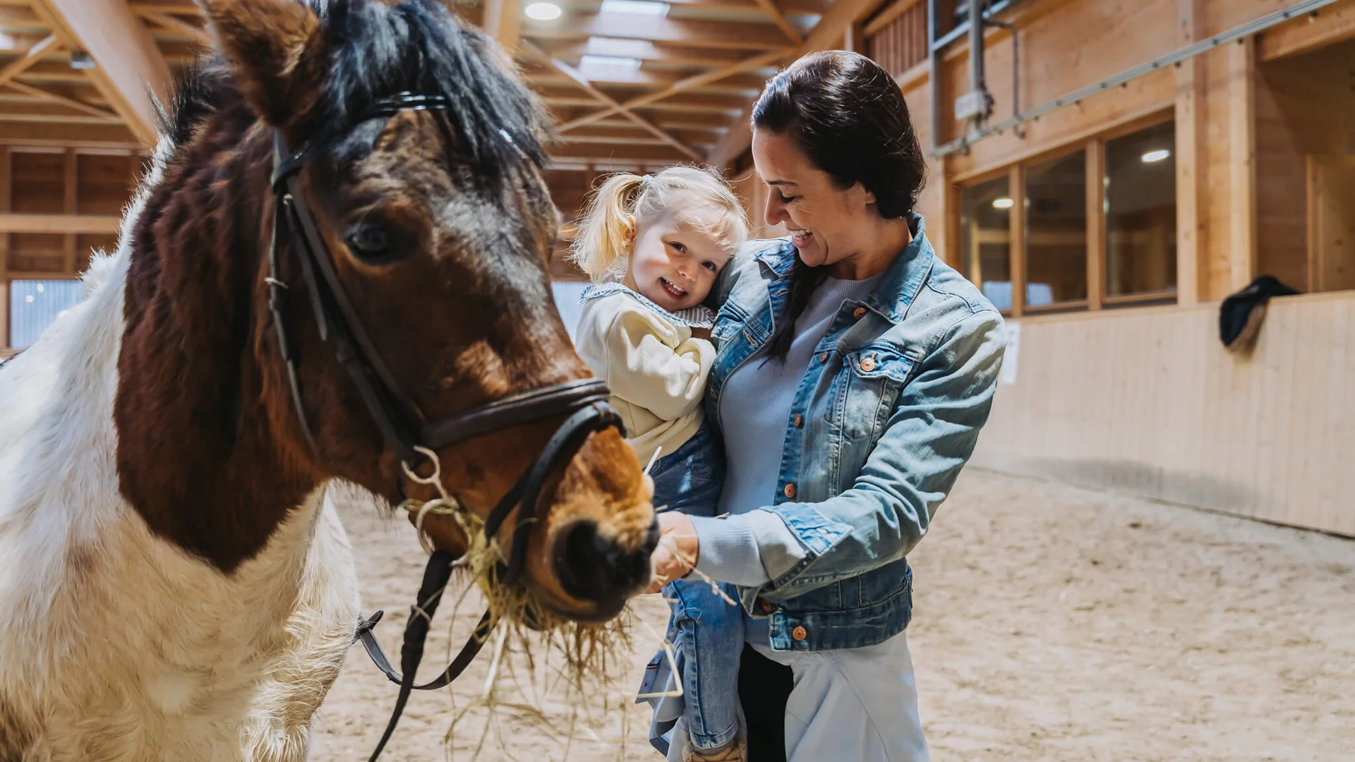 Familienzeit im Kinderhotel Buchau am Achensee Frau hält lachendes Mädchen im Innenstall neben fressendem Pony
