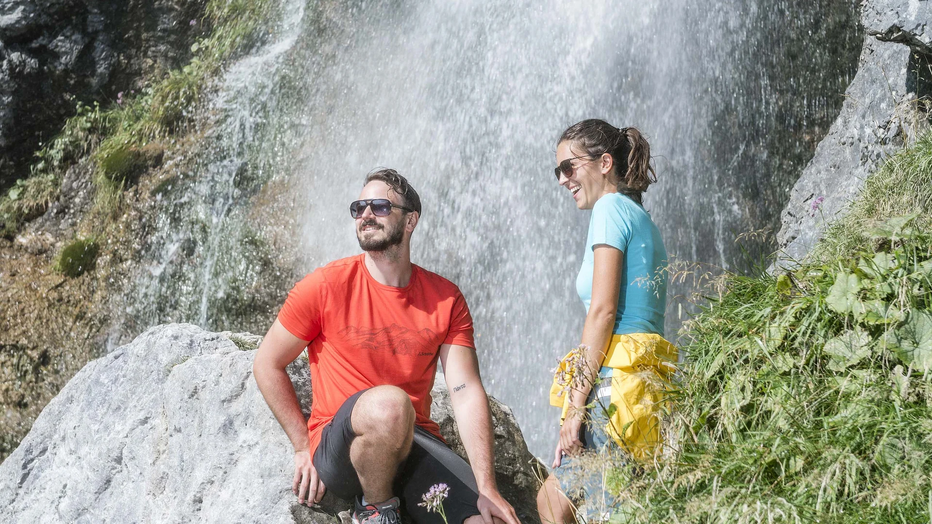 Vom Wanderhotel in Tirol direkt in die Natur Mann in rotem Shirt und Frau in blauem Shirt sitzen neben Wasserfall auf Felsen
