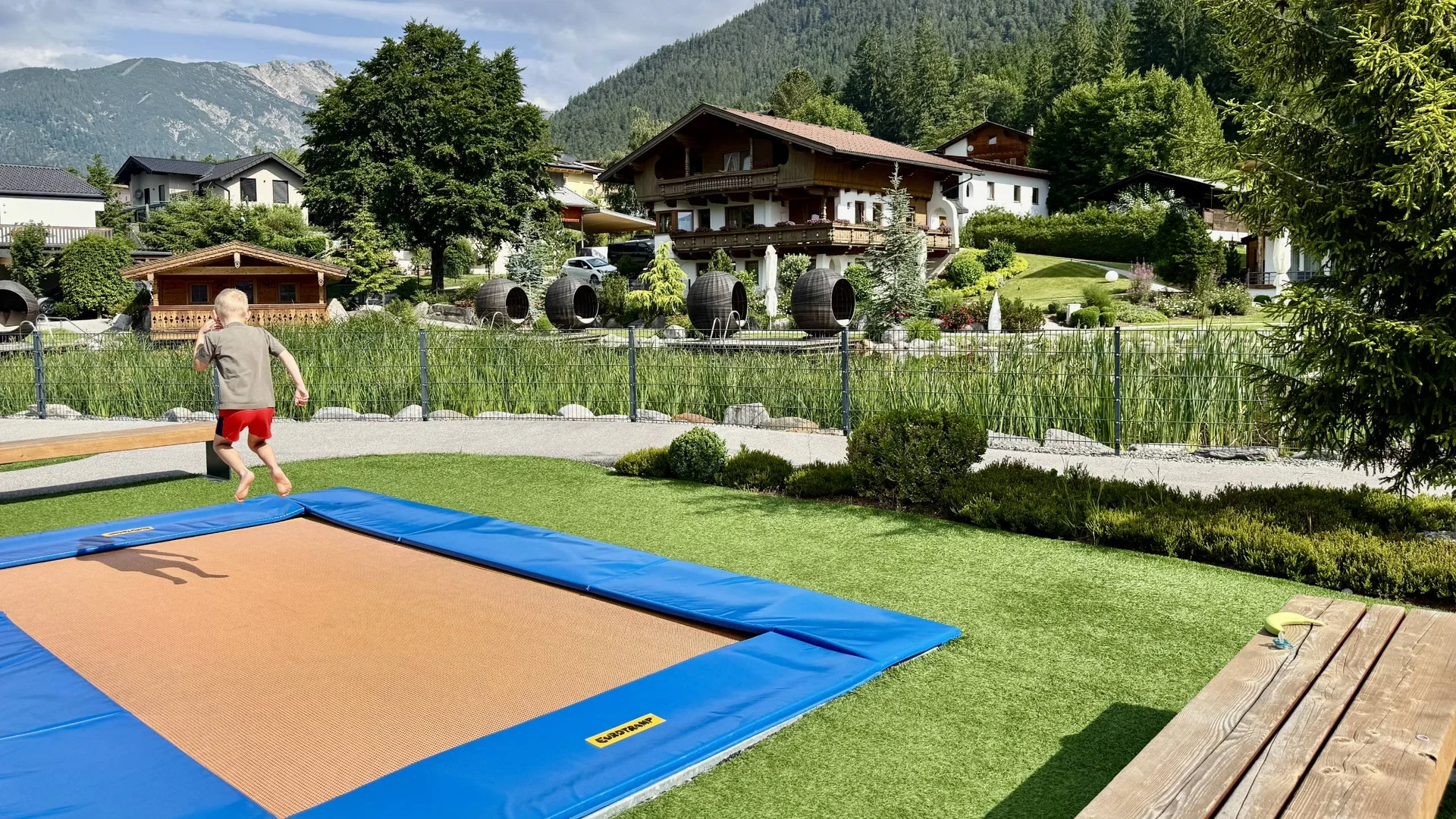 Outdoorspaß im Familienhotel am See in Österreich Kind springt auf Trampolin in bergigem Garten vor traditionellen Häusern
