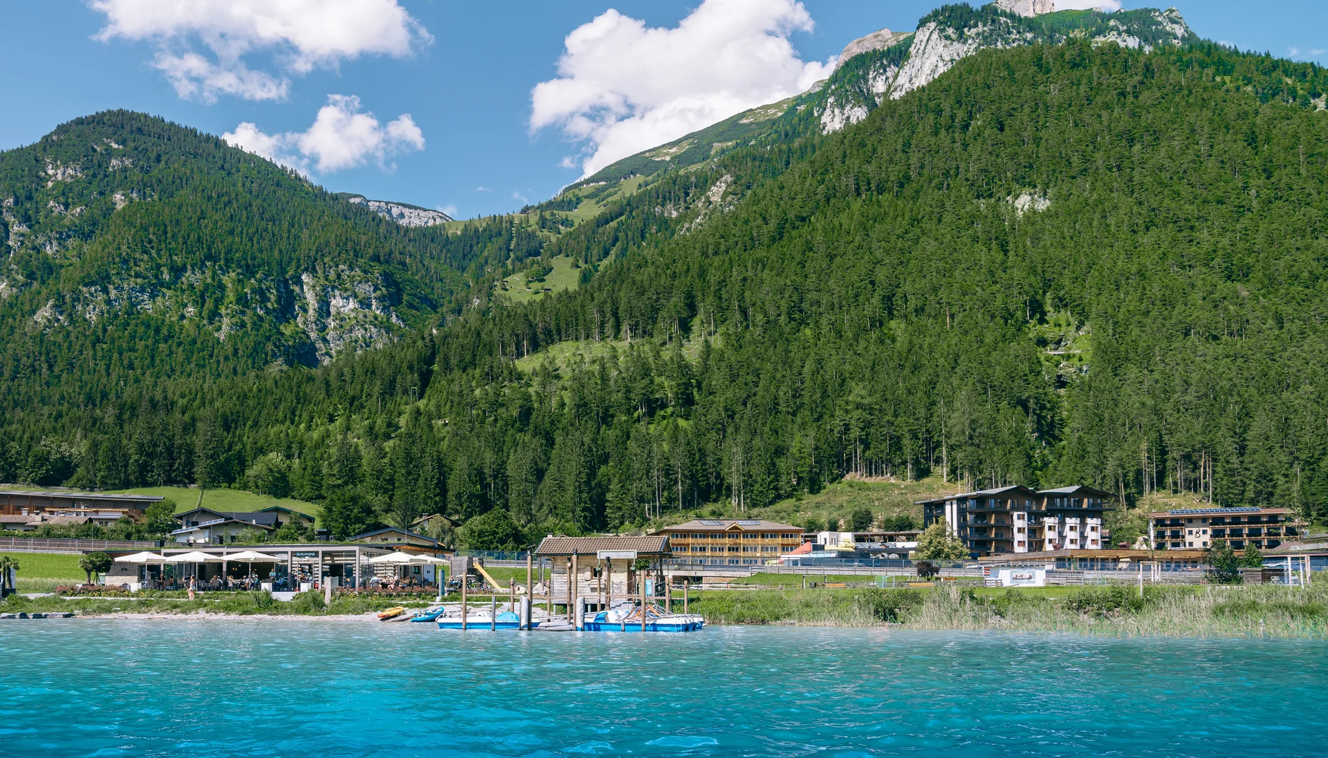 Entspannte Ferien für die ganze Familie. Berge und Wald hinter einem See mit Häusern und Steg am Ufer unter blauem Himmel