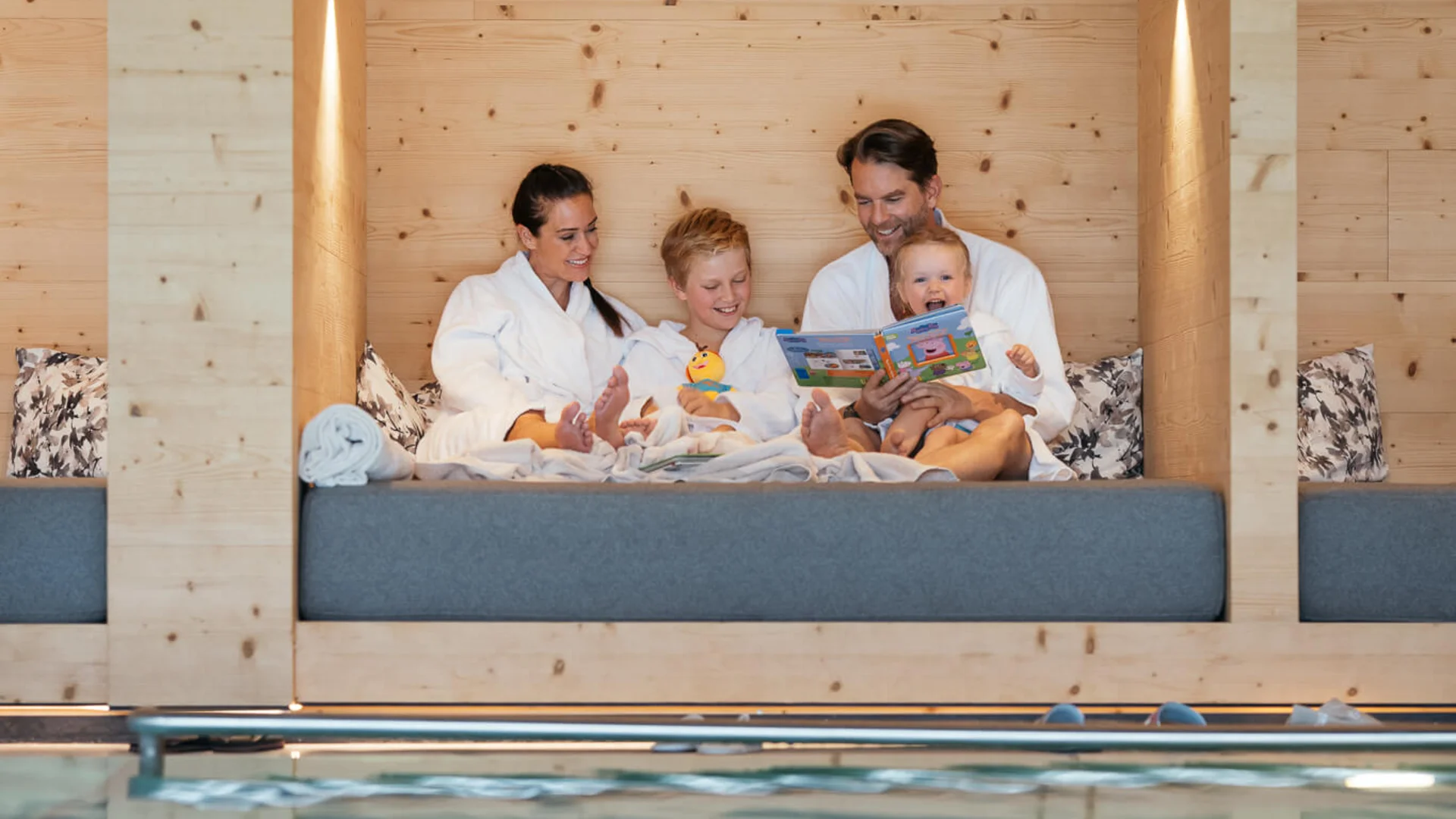 Wasserspaß im Familienhotel in Tirol mit Pool Familie in Bademänteln sitzt gemütlich an einem Holzruheraum neben dem Pool