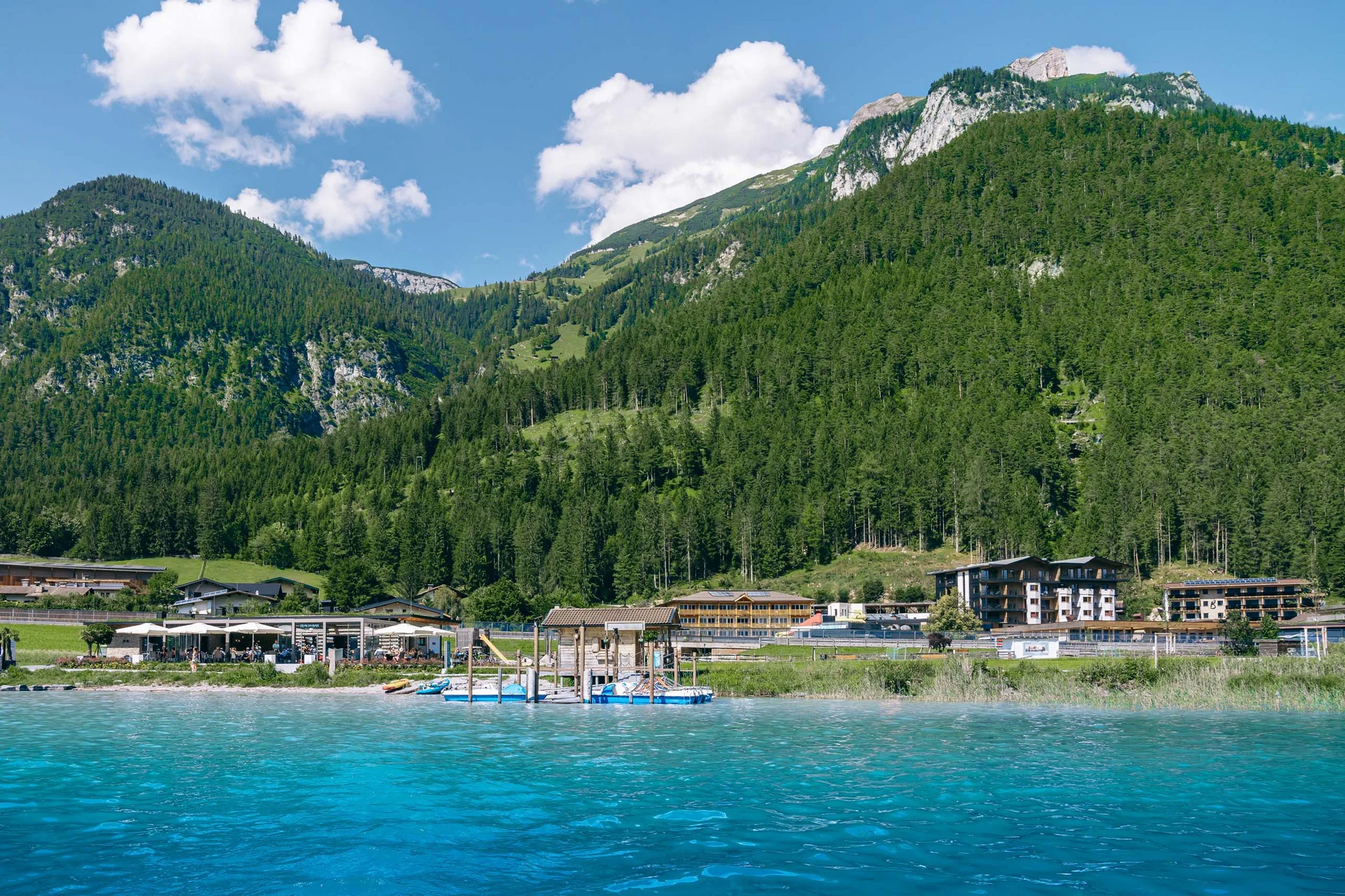 Willkommen im Familienhotel Buchau am Achensee Klares blaues Wasser mit Bergkulisse und Gebäude am Ufer bei sonnigem Wetter