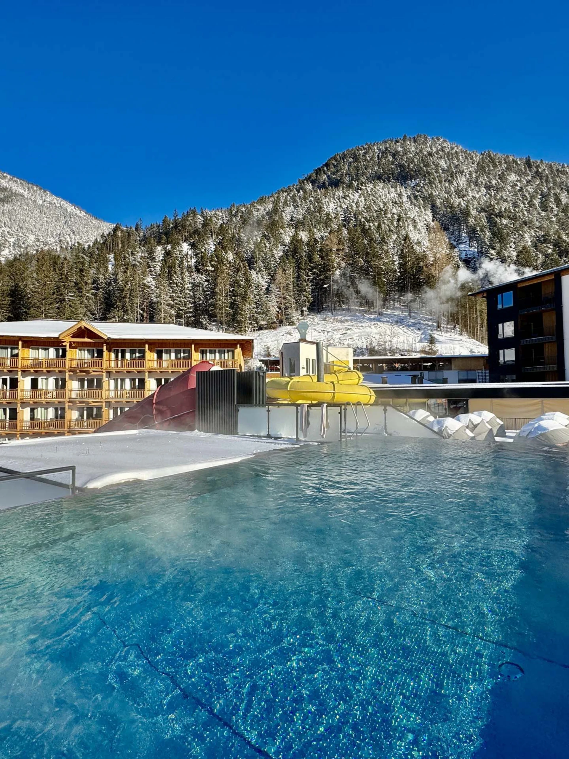 Wir lassen Bilder sprechen. Beheizter Außenpool mit Wasserrutsche in verschneiter Berglandschaft bei klarem Himmel