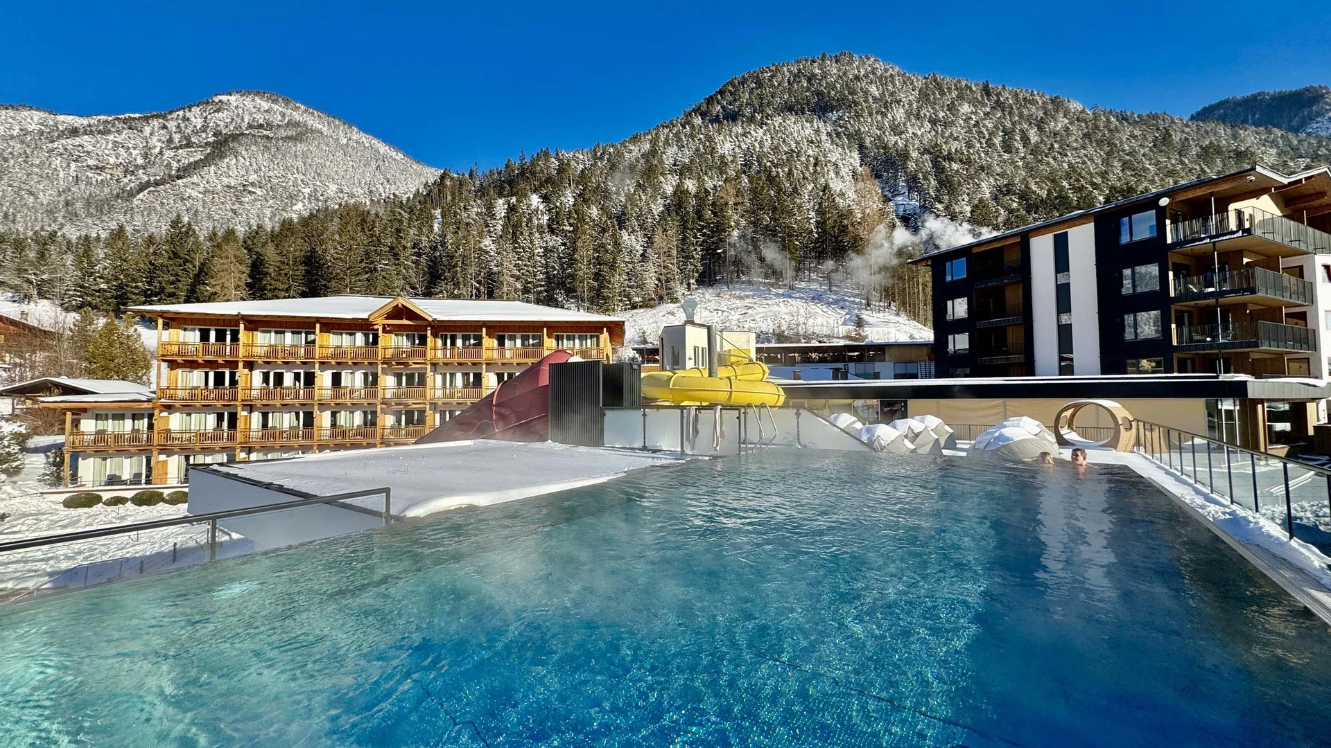 Willkommen im Familienhotel Buchau am Achensee Beheizter Außenpool mit Wasserrutsche in verschneiter Berglandschaft bei klarem Himmel