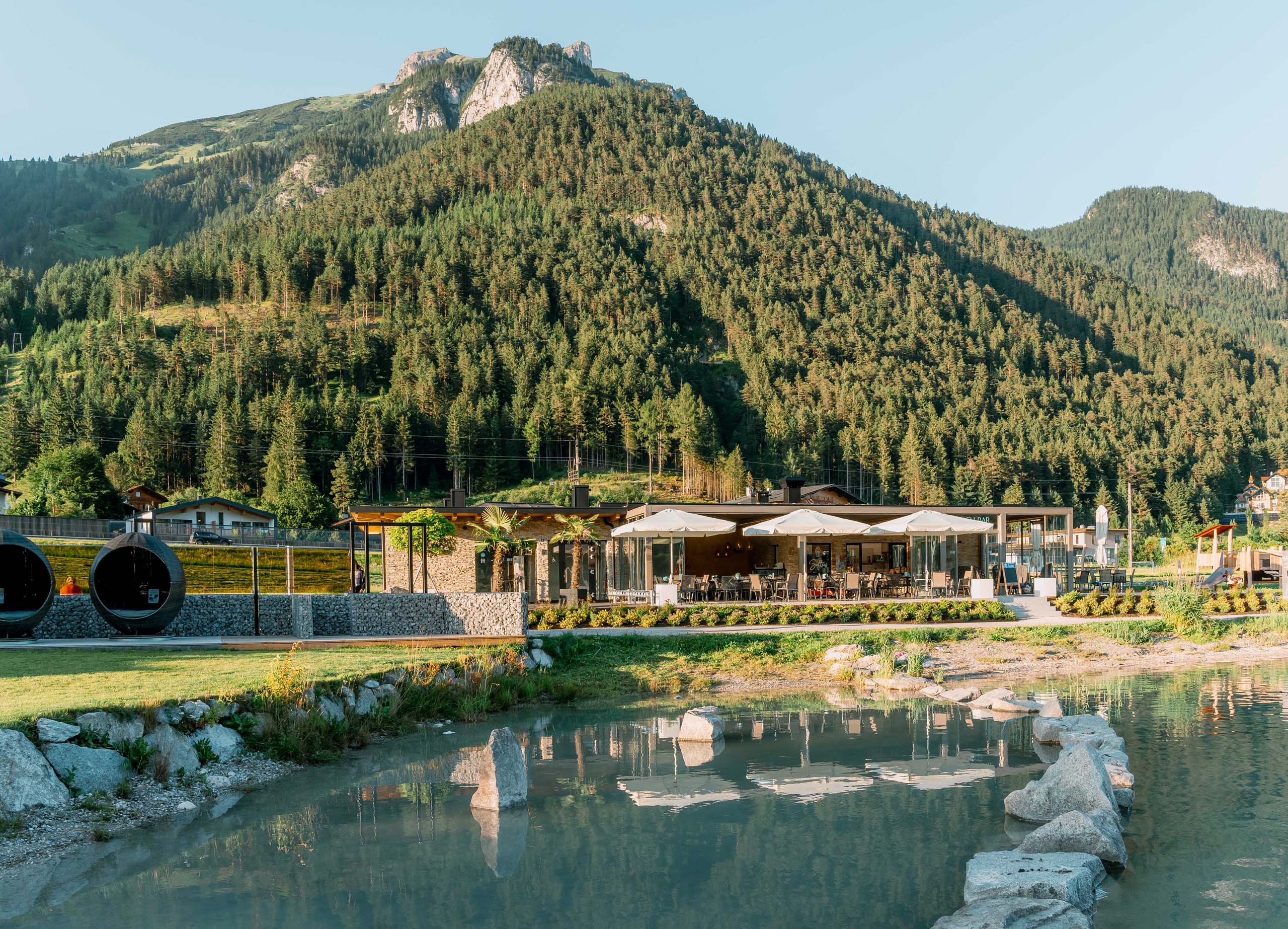 Wir lassen Bilder sprechen. Restaurant mit Sonnenschirmen am See vor bewaldetem Berg unter klarem Himmel