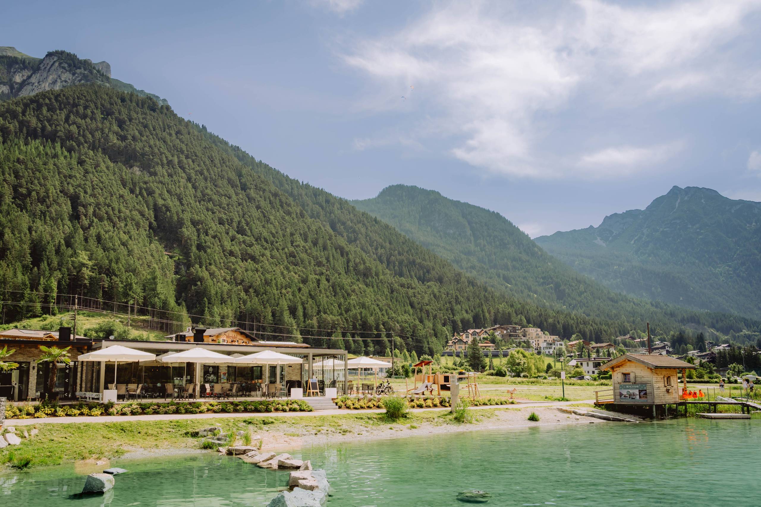 Wir lassen Bilder sprechen. See mit Restaurant, Spielplatz und Berghütten vor bewaldeten Bergen