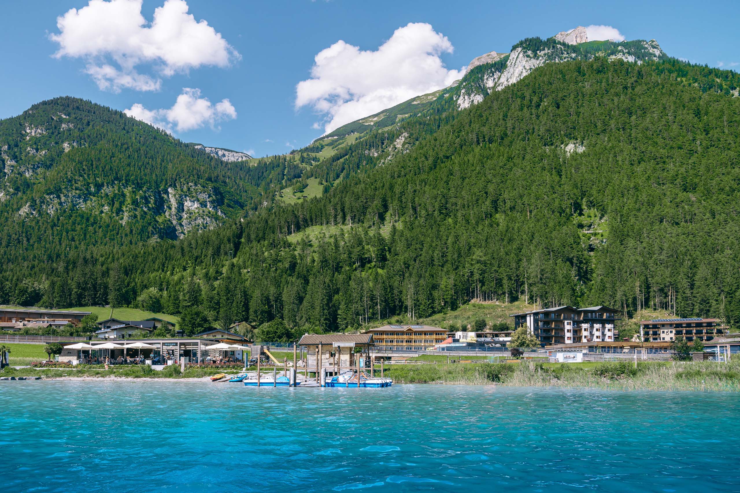 Wir lassen Bilder sprechen. Klares blaues Wasser mit Bergkulisse und Gebäude am Ufer bei sonnigem Wetter