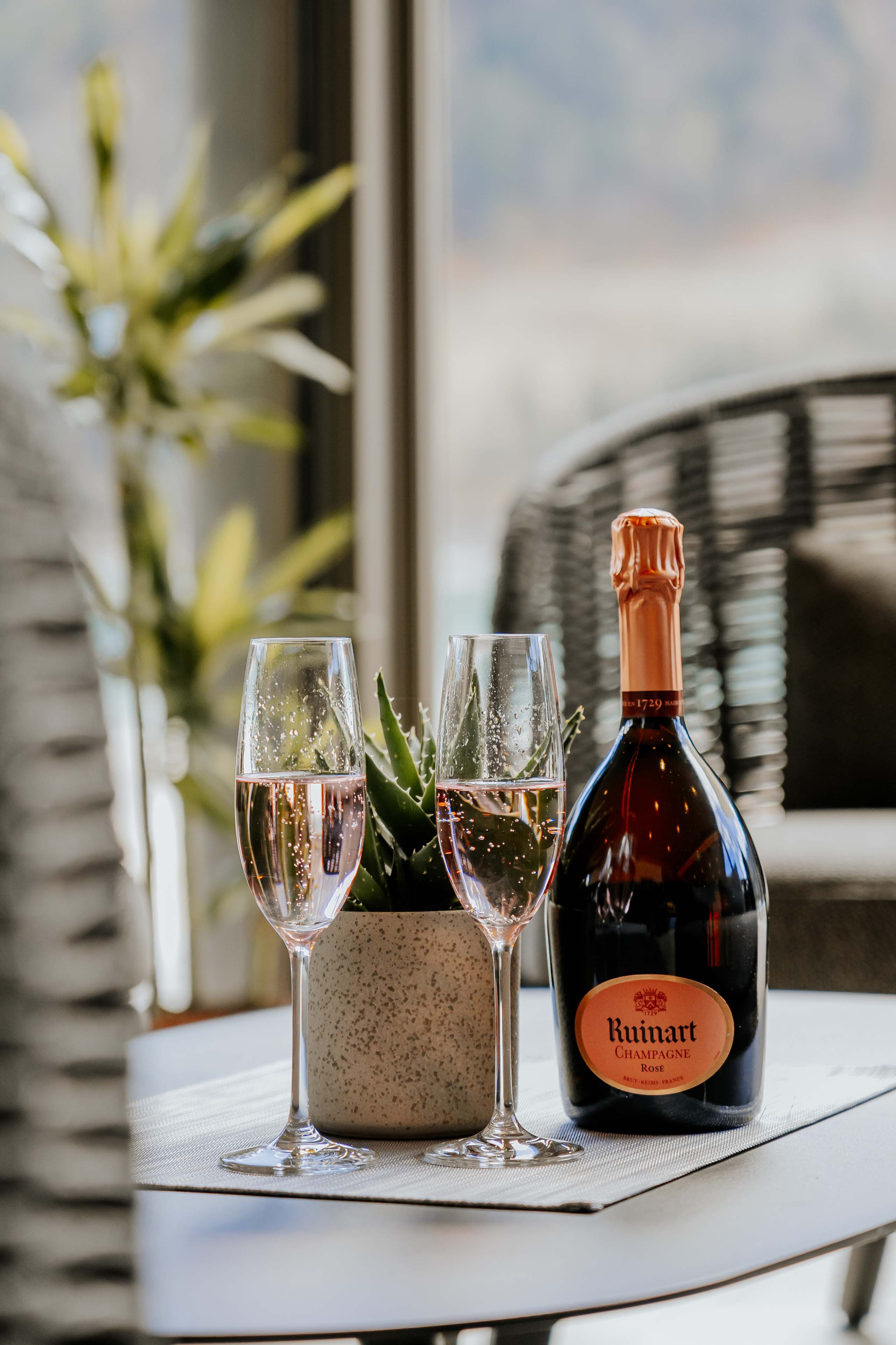 Wir lassen Bilder sprechen. Zwei Champagnergläser und eine Flasche Ruinart Rosé auf einem Tisch mit einer kleinen Pflanze