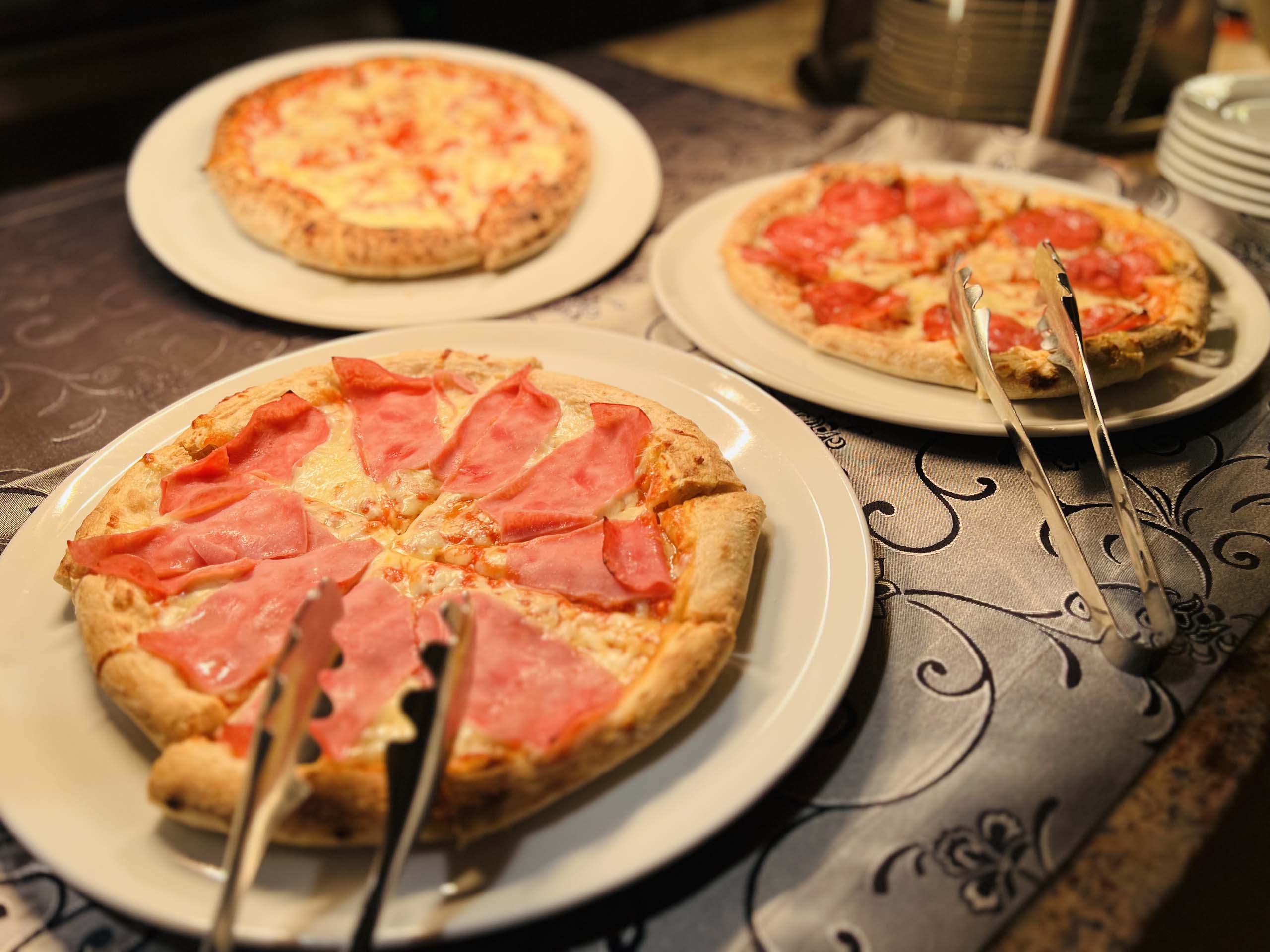 Wir lassen Bilder sprechen. Drei verschiedene Pizzen auf Tellern auf einem Tisch mit Servierzangen