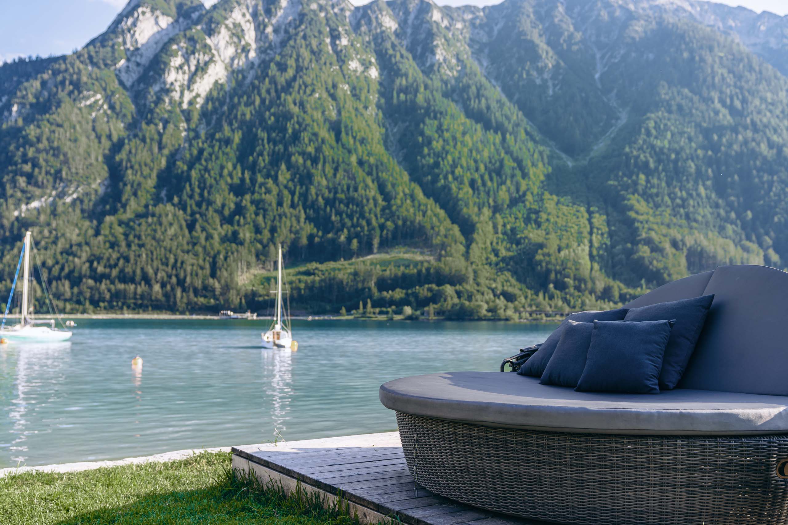Wir lassen Bilder sprechen. Rundes Lounge-Sofa mit Kissen am See mit Segelbooten und bewaldeten Bergen im Hintergrund