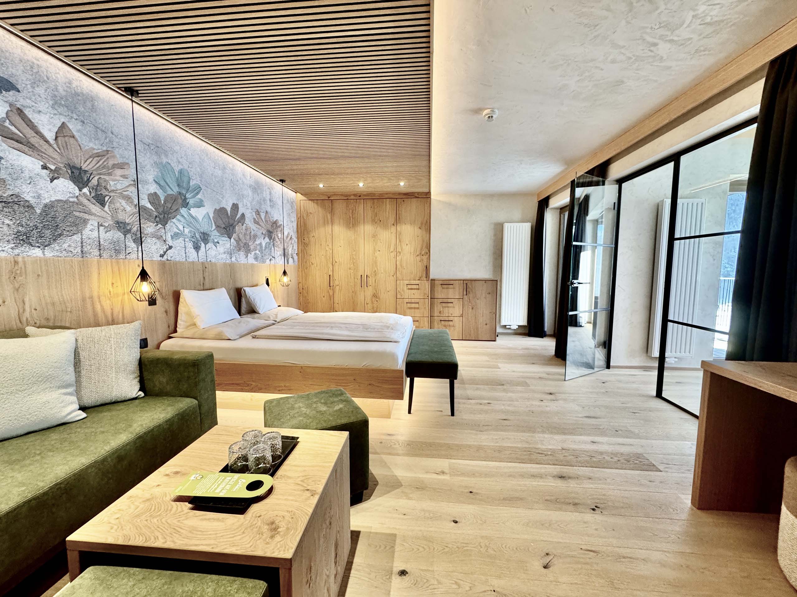 Wir lassen Bilder sprechen. Modernes Hotelzimmer mit Holzboden, grünem Sofa und Doppelbett mit Blumen-Wandbild