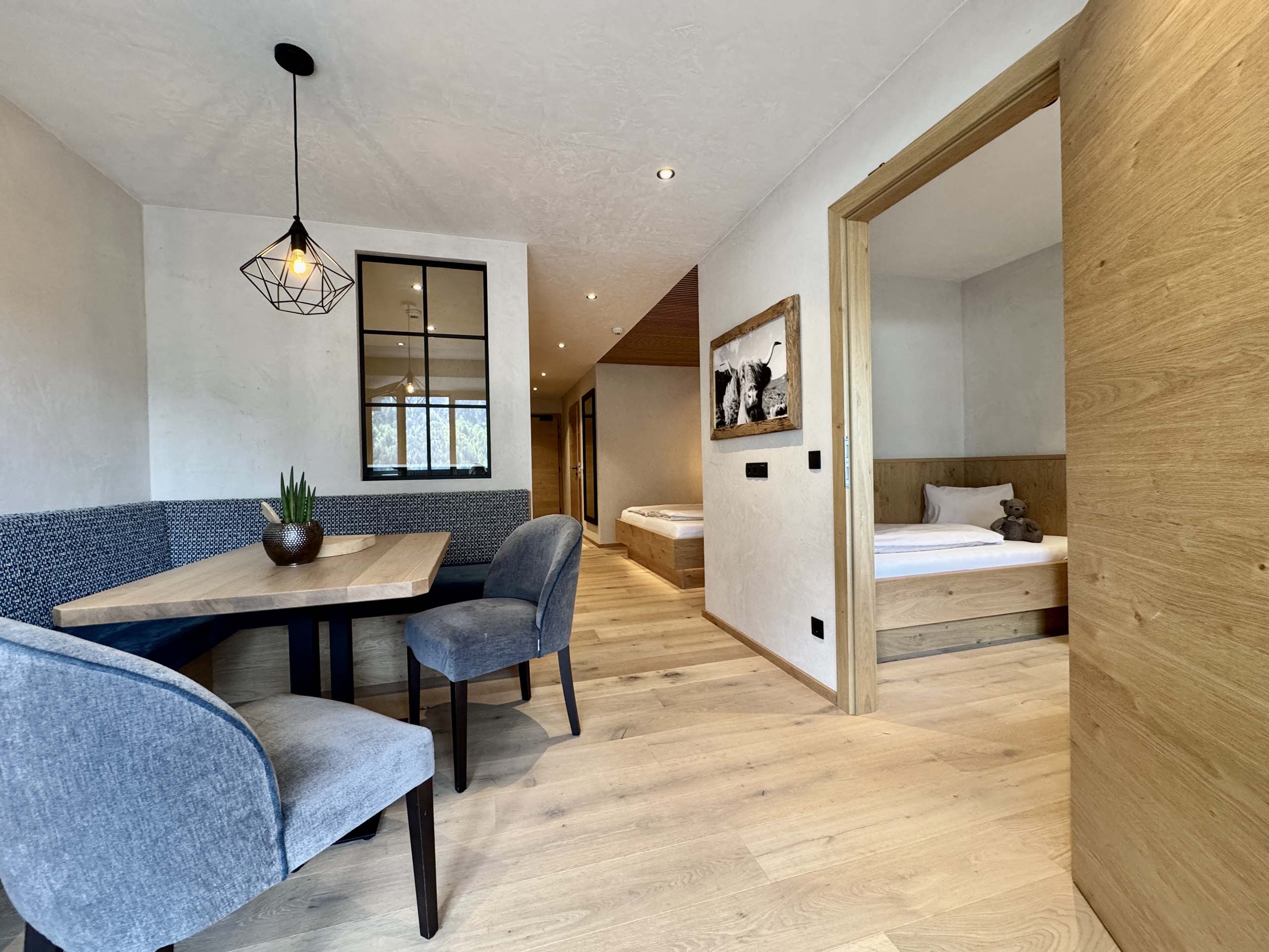 Wir lassen Bilder sprechen. Modernes Hotelzimmer mit Holzboden, Tisch mit Stühlen und Blick ins Schlafzimmer