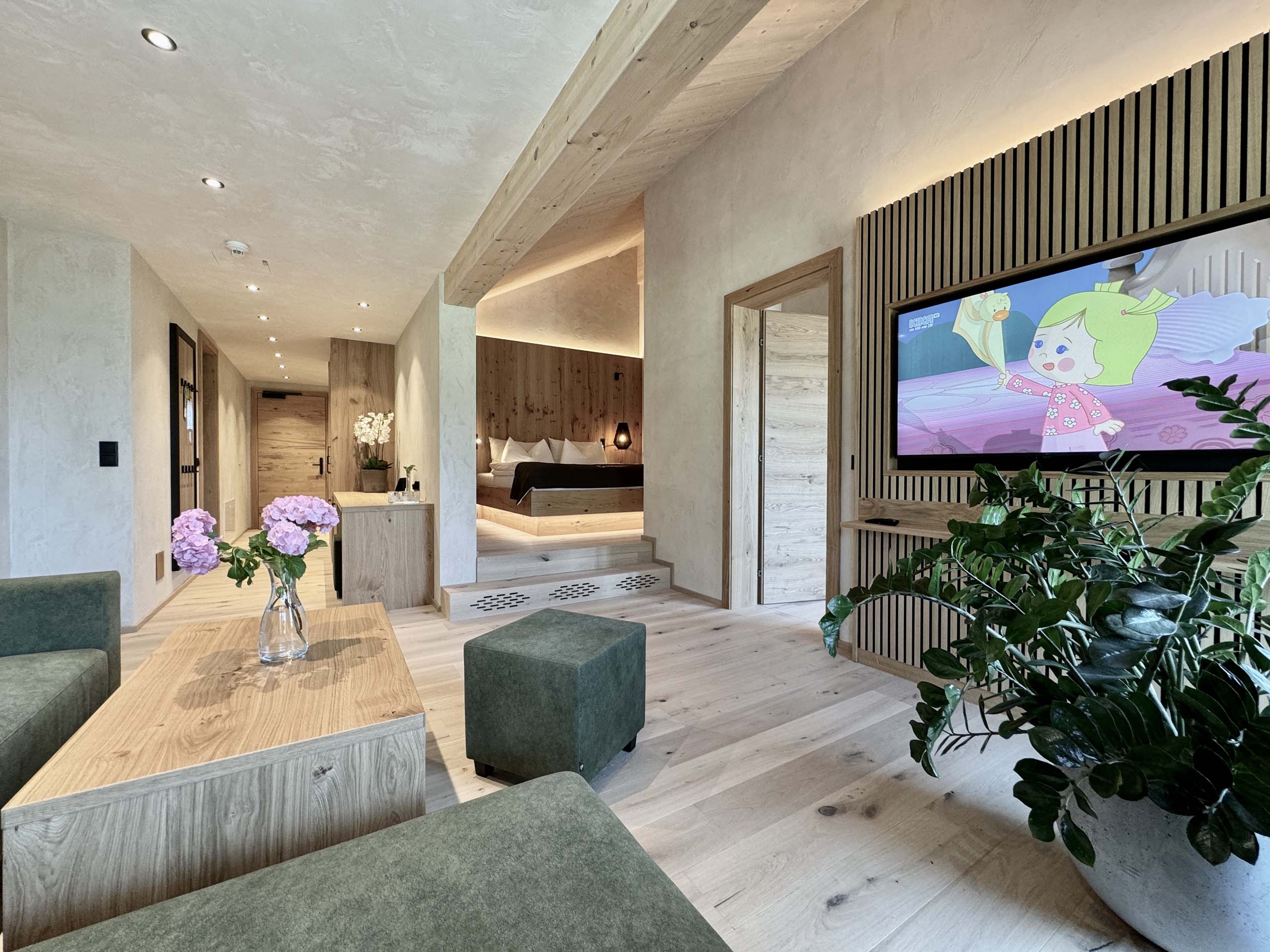 Wir lassen Bilder sprechen. Moderner Hotelwohnraum mit grünem Sofa, TV und Schlafzimmer im Hintergrund