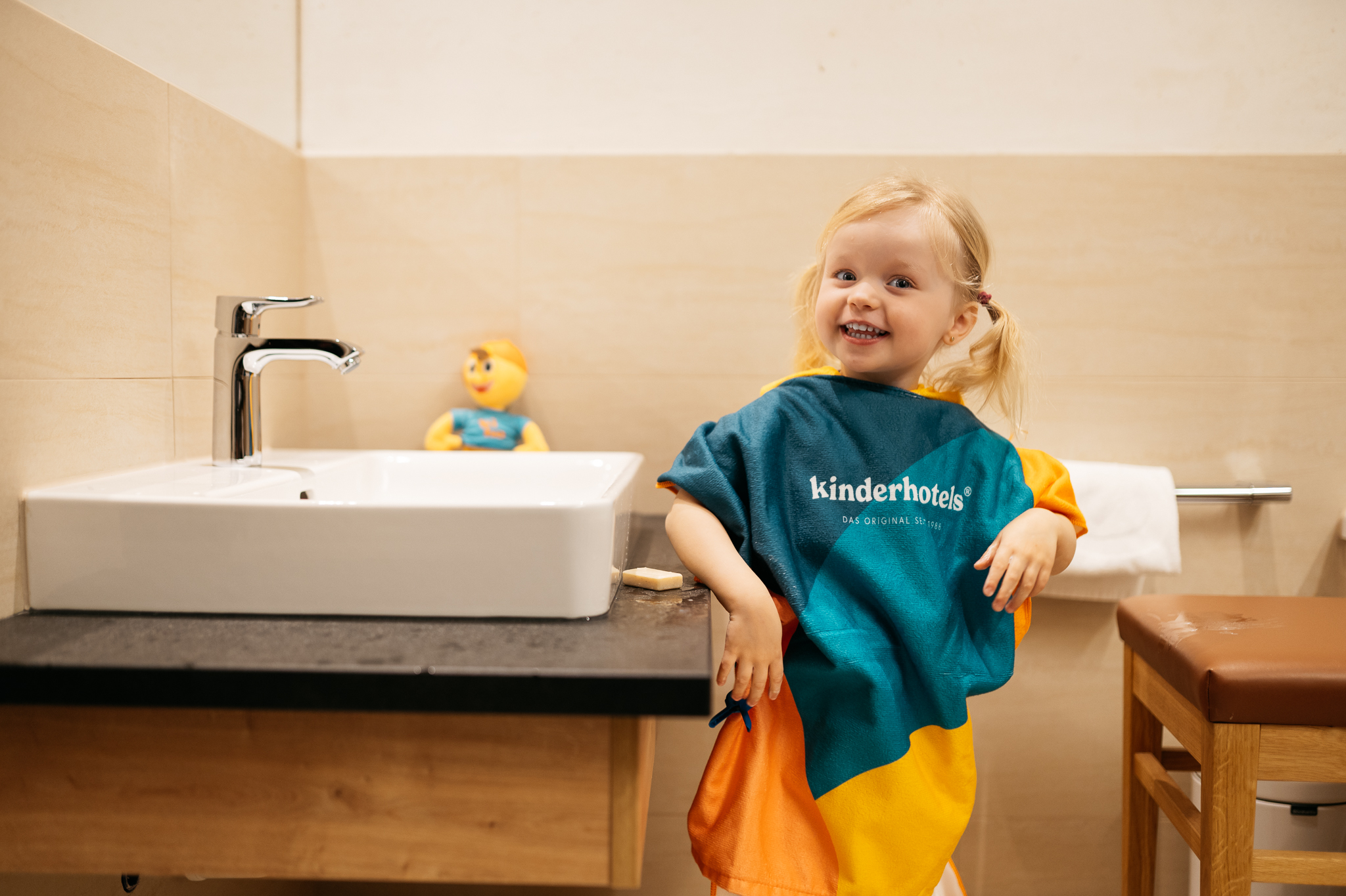 Wir lassen Bilder sprechen. Fröhliches Mädchen mit Kinderhotel Poncho im Badezimmer neben Waschbecken