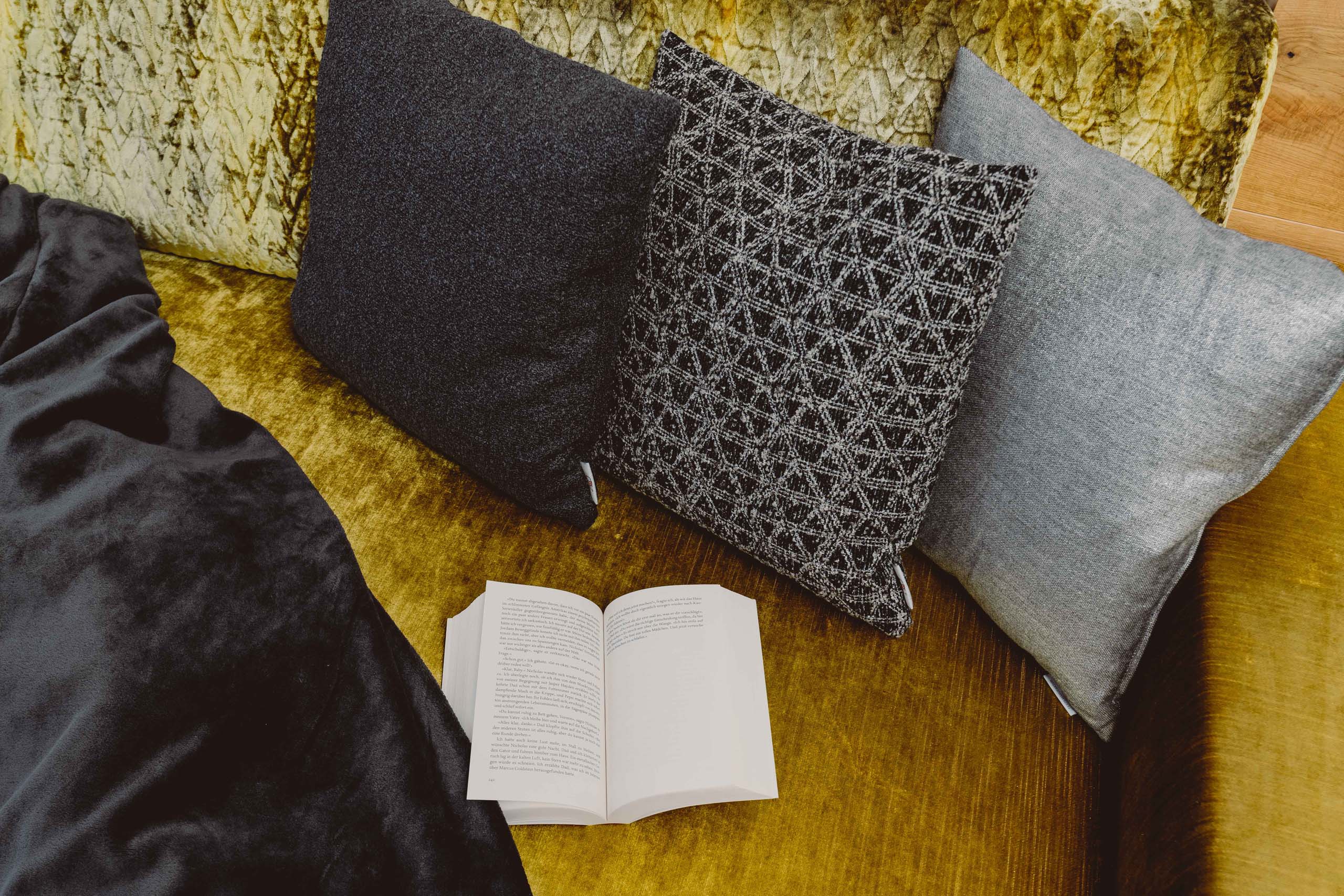 Wir lassen Bilder sprechen. Gemütliche Sitzecke mit drei Kissen, offenes Buch und Decke auf gelbem Sofa