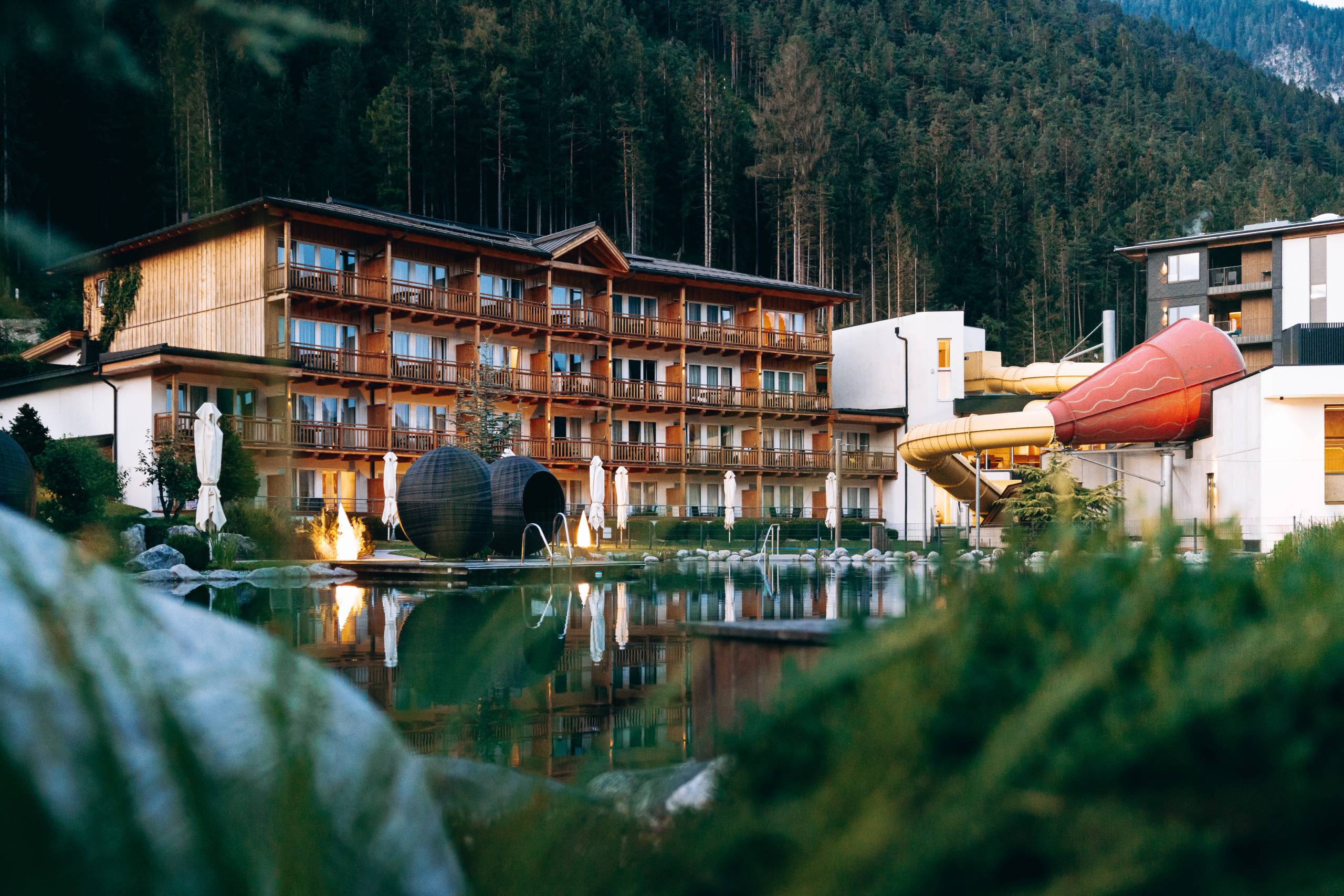 Wir lassen Bilder sprechen. Hotel mit Holzfassade und Wasserrutsche neben einem See vor Waldkulisse